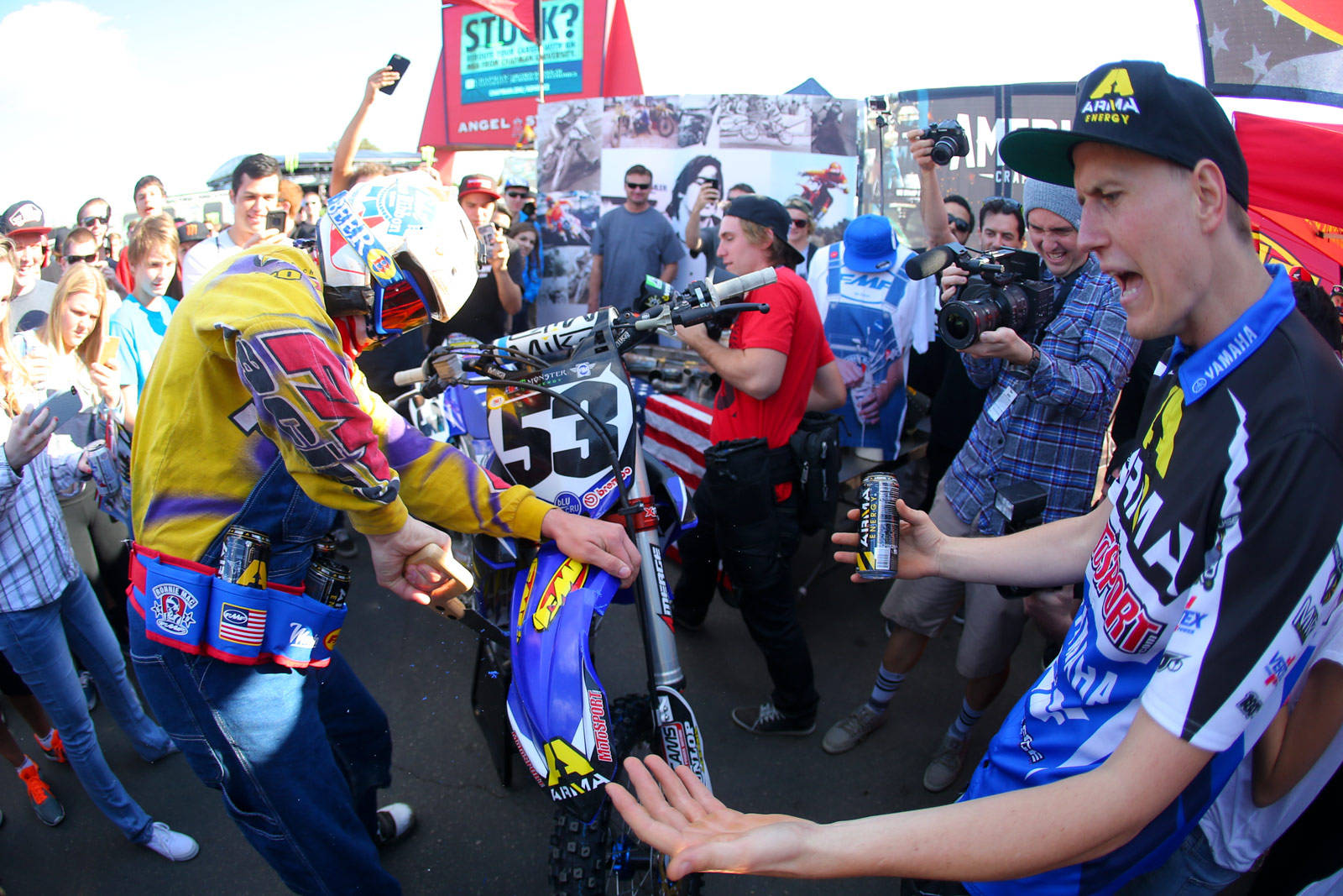 Ronnie Mac 桌布 壁紙 — 兩名身穿色彩繽紛裝備的越野摩托車手在藍色摩托車旁握手，粉絲和攝影師在陽光明媚的戶外活動中圍繞著他們。