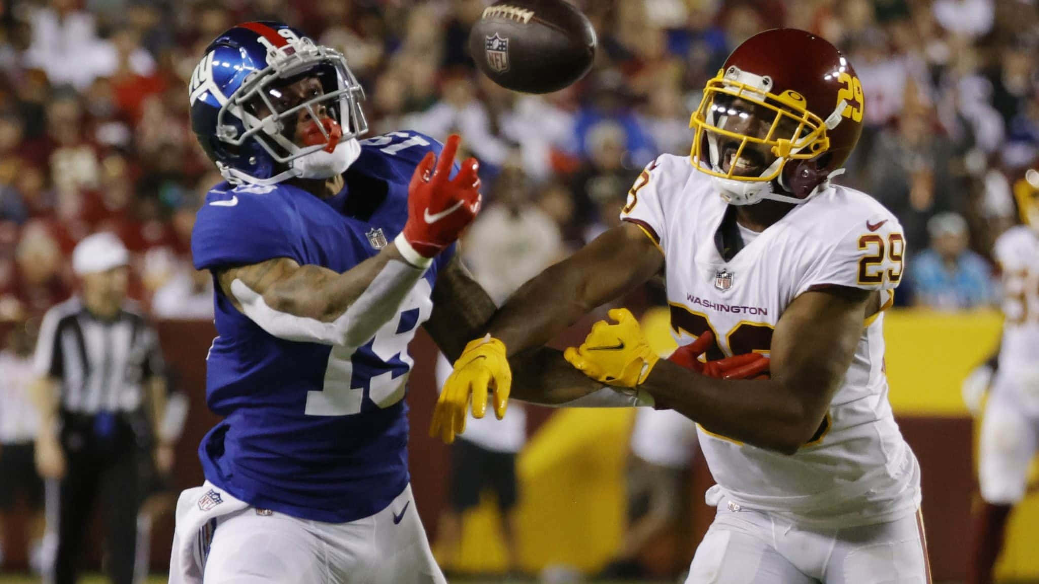 足球 桌布 壁紙 — 兩名身穿藍白球衣的NFL球員在夜間比賽中，於球場燈光下在空中爭奪橄欖球。