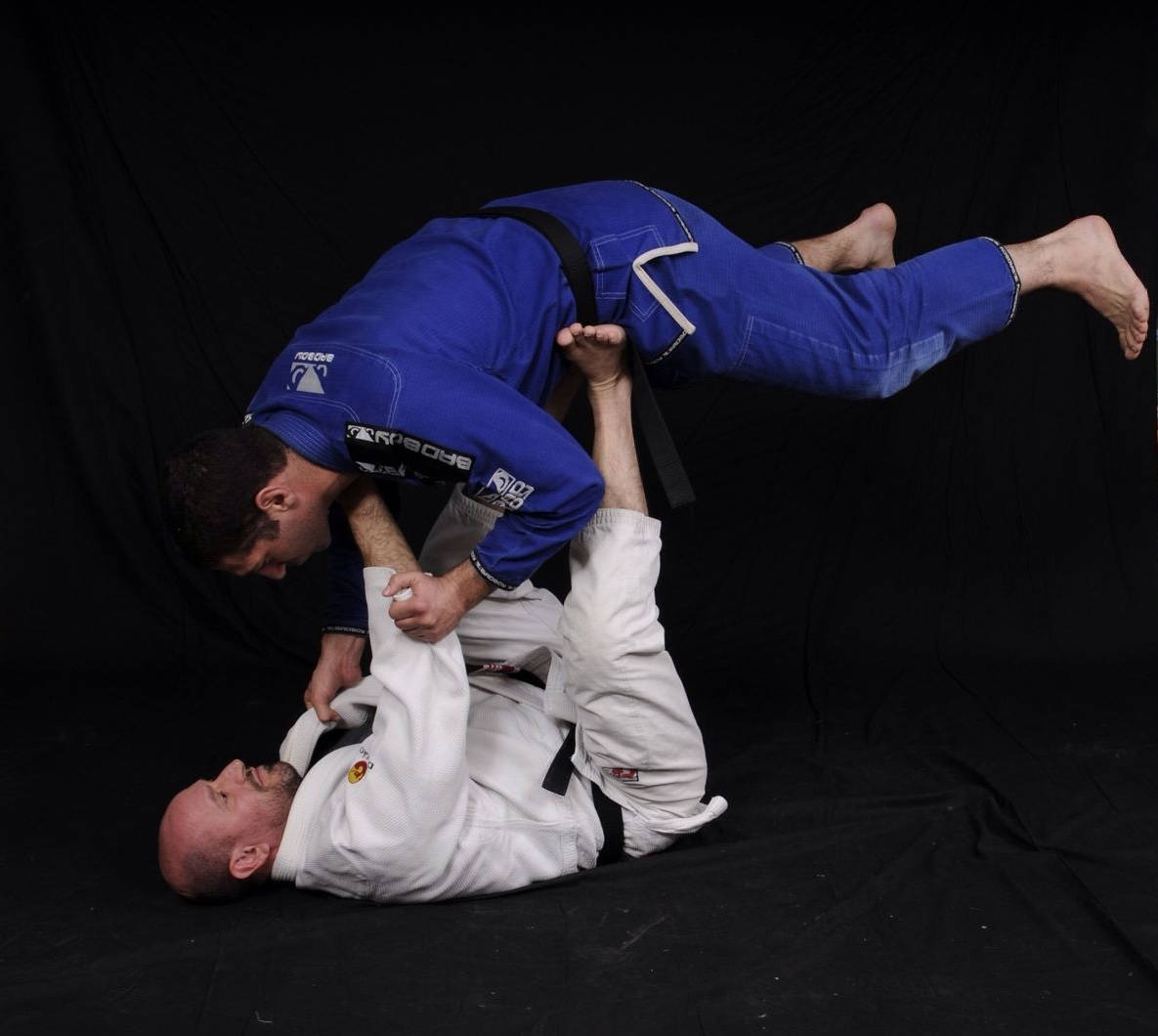 martial arts 桌布 壁紙 — 兩名身穿藍白色柔道服的男人在純黑背景下執行一個動態的巴西柔術降服技。