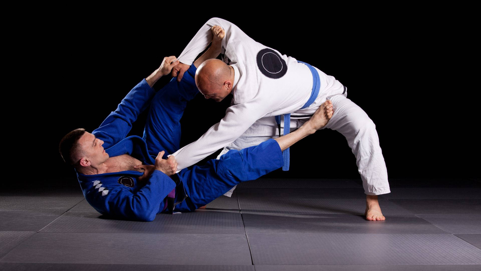martial arts 桌布 壁紙 — 兩名身穿藍白色柔道服的男性柔道運動員在黑色背景的深色墊子上執行一個動態的纏鬥動作。