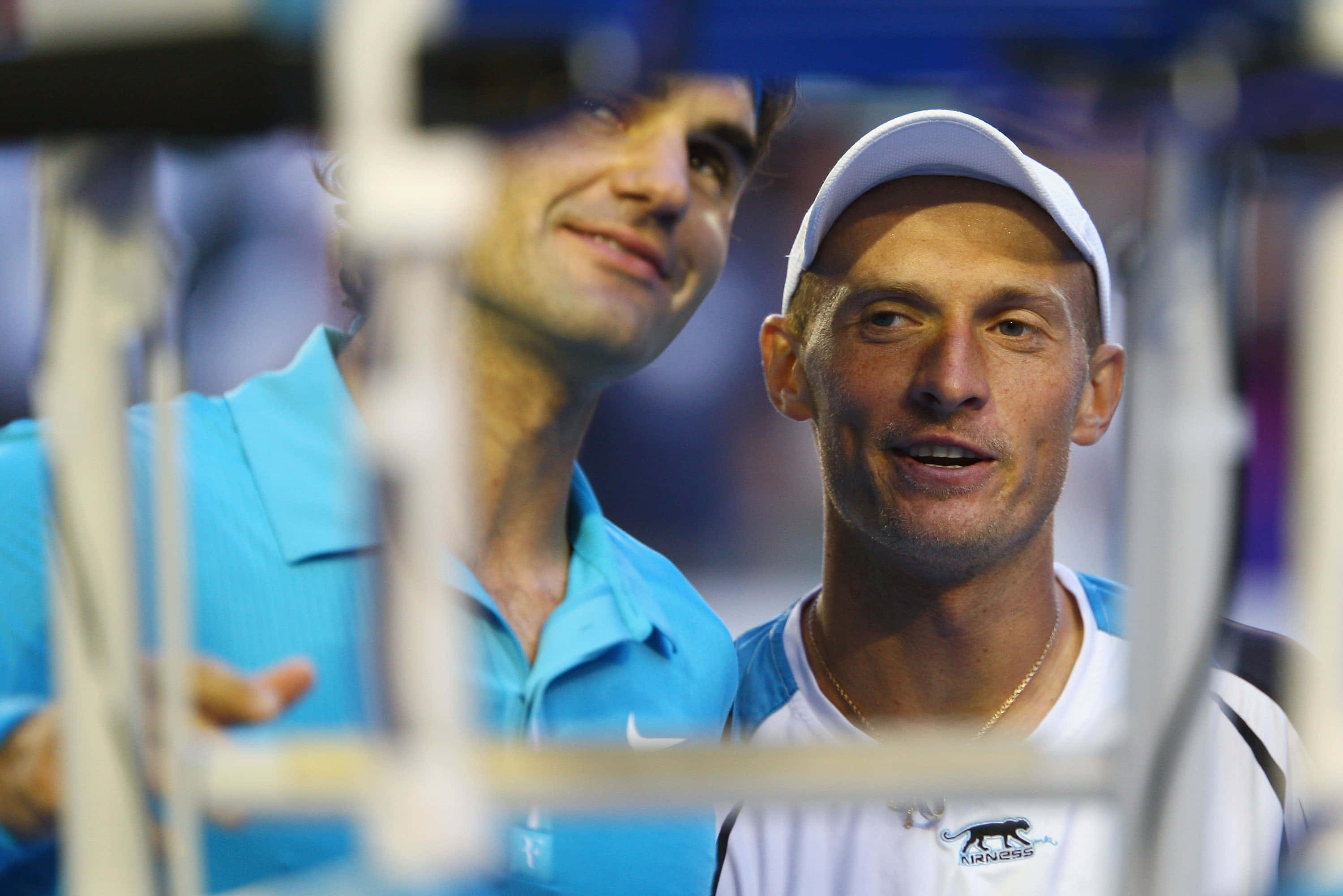 Tennis 桌布 壁紙 — 兩名身穿藍白裝備、面帶微笑的男網球選手在比賽期間透過模糊的網障凝視。
