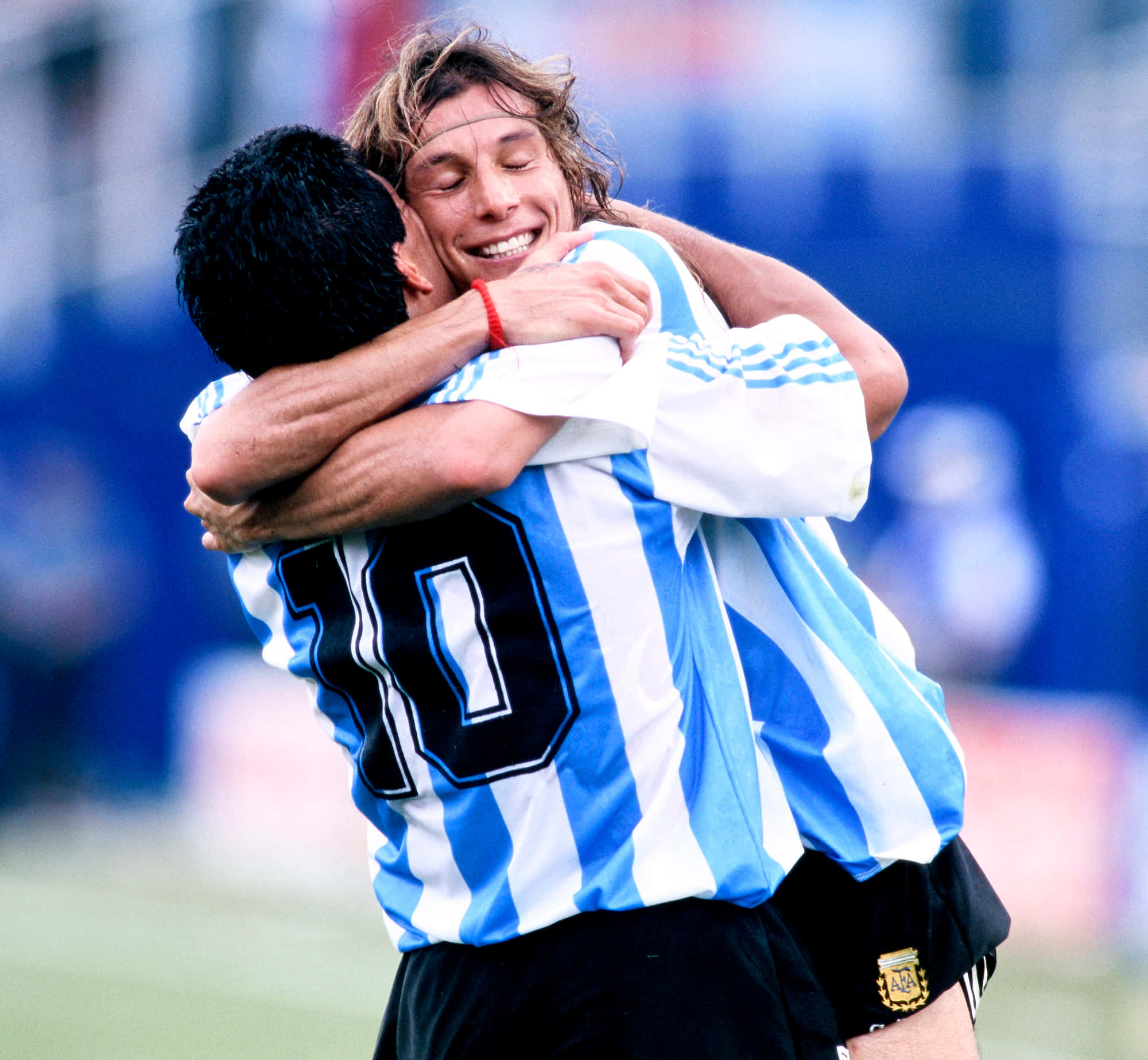 Argentina National Football Team 桌布 壁紙 — 兩名身穿藍白間條球衣的阿根廷足球員在綠色球場上愉快地擁抱，背景的體育館座位模糊不清。