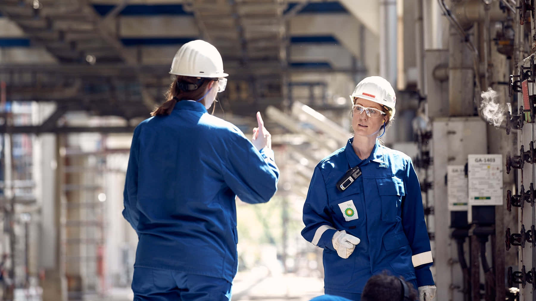 engineering 桌布 壁紙 — 兩名身穿藍色制服和白色安全帽的工程師在明亮的日光下，在工業管道和機械之間交談。