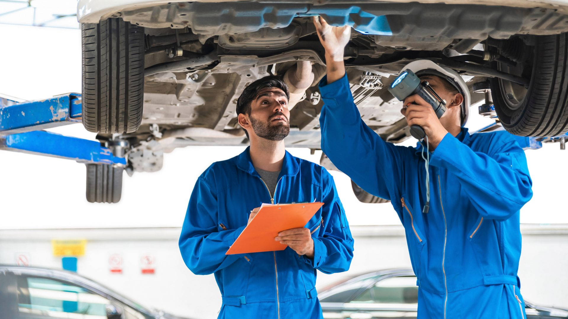 engineering 桌布 壁紙 — 兩名身穿藍色制服的機械師在明亮的車庫中用工具和掃描器檢查一輛升起汽車的底盤。