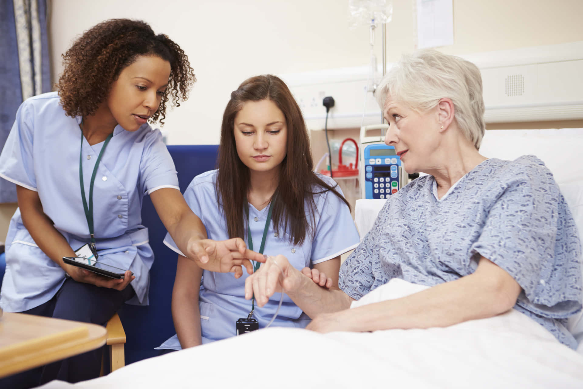 Nursing 桌布 壁紙 — 兩名身穿藍色工作服的護士在醫院病床上協助一位老年病人使用醫療設備。