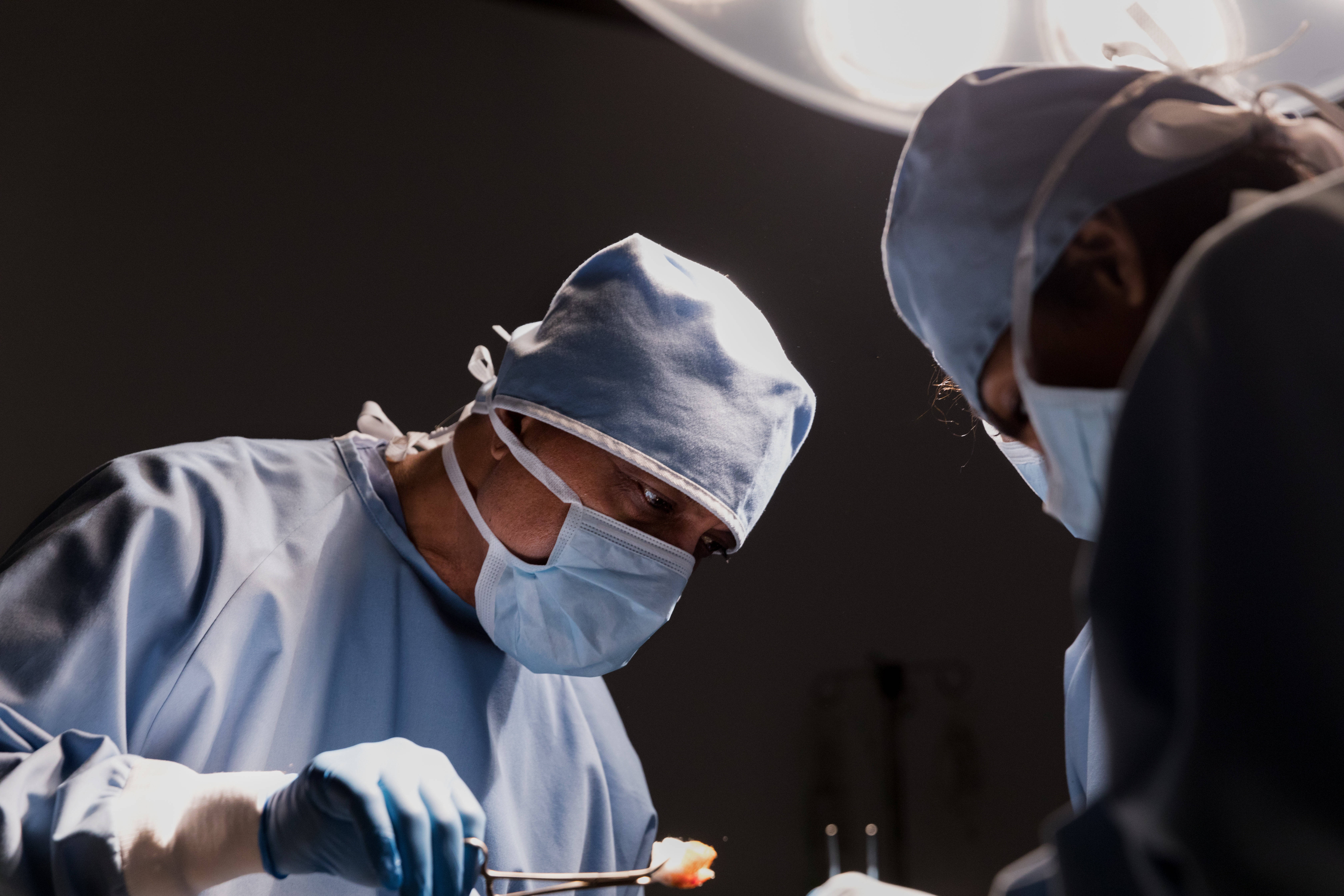 Doctors 桌布 壁紙 — 兩名身穿藍色手術服和口罩的外科醫生在明亮的手術燈下專注地進行精確的手術。
