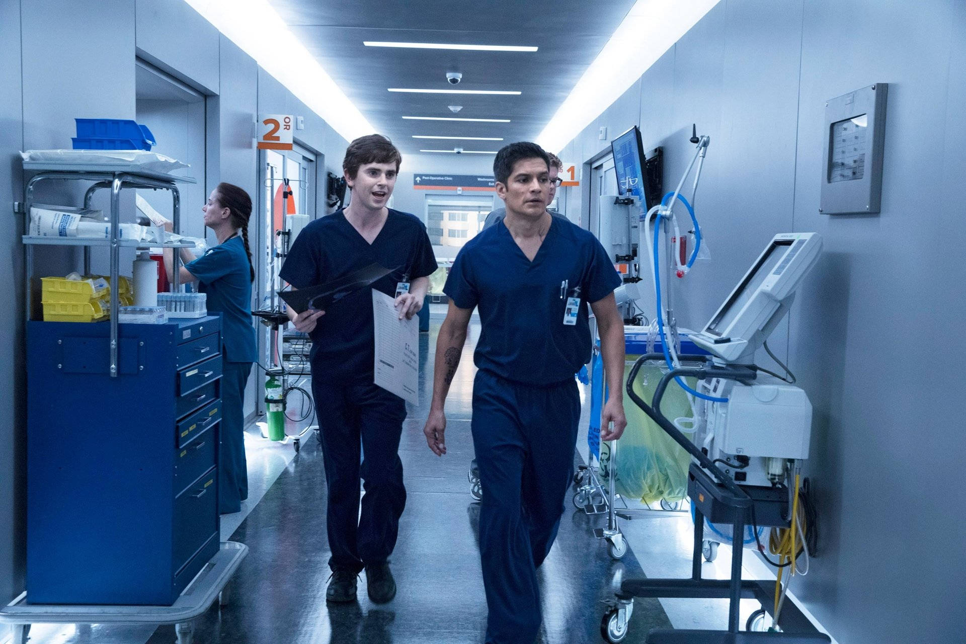 The Good Doctor 桌布 壁紙 — 兩名身穿藍色手術服的男醫生快步走過無菌的醫院走廊，經過醫療設備和一名護士。