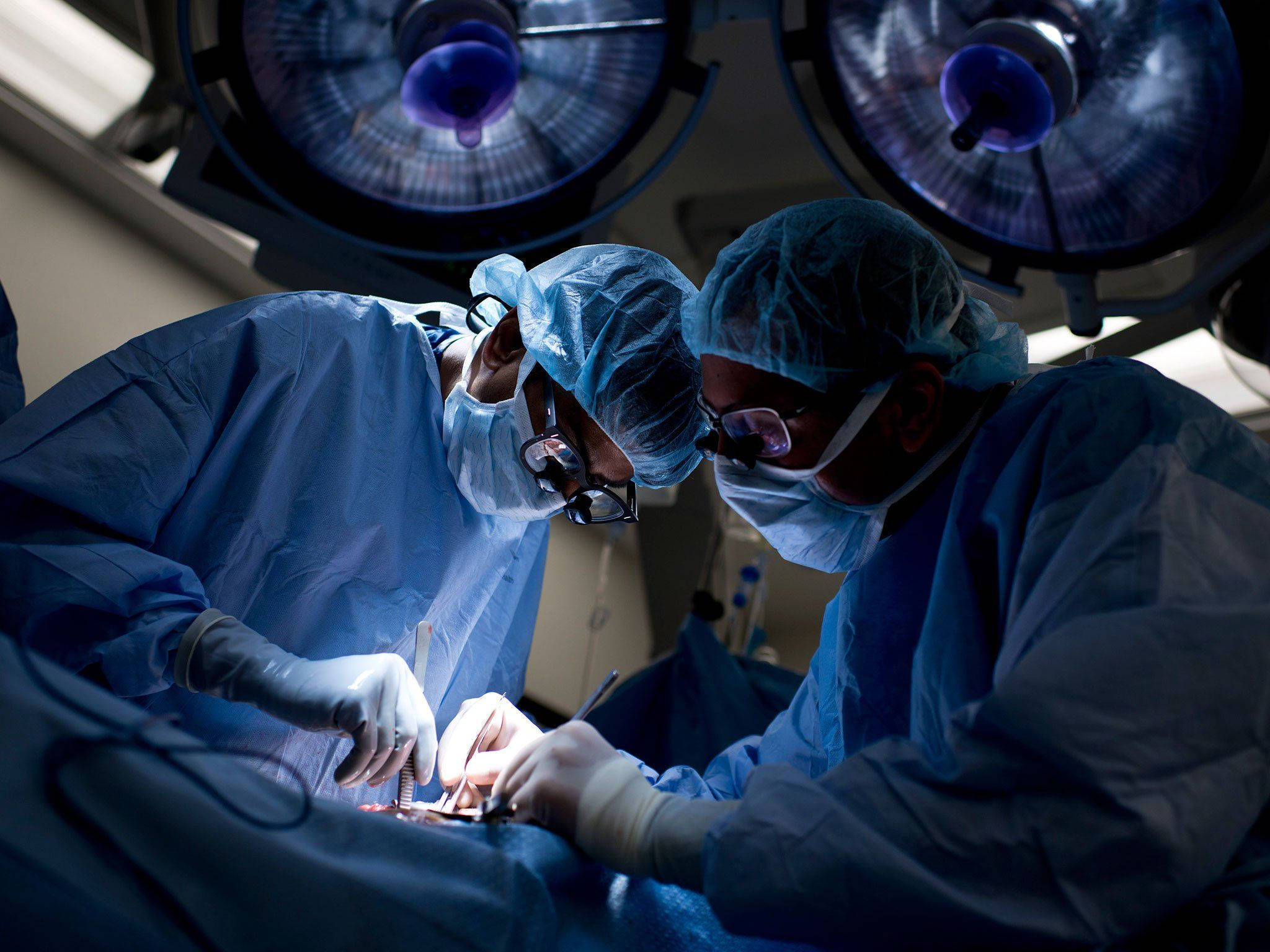 Surgeon 桌布 壁紙 — 兩名身穿藍色手術衣和口罩的外科醫生在明亮的手術燈下，在無菌手術室中進行精細的手術。