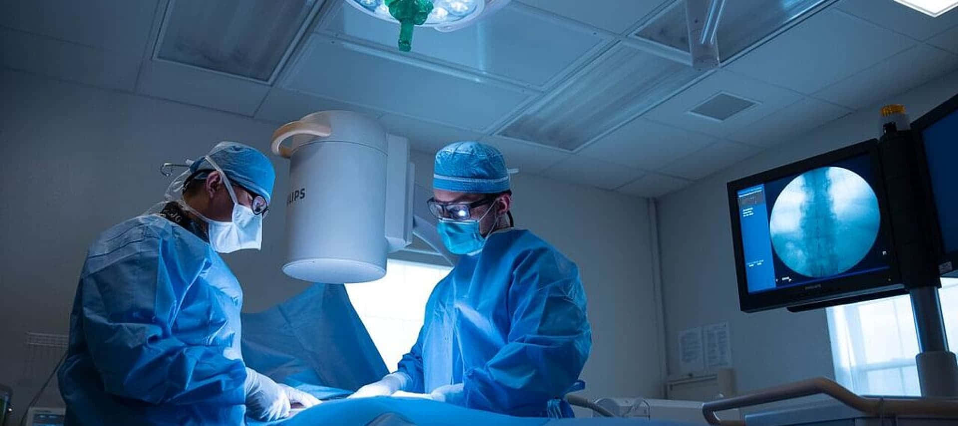 醫生 桌布 壁紙 — 兩名身穿藍色手術衣和口罩的外科醫生在無菌手術室中，於明亮的手術燈下專注地為病人進行手術。