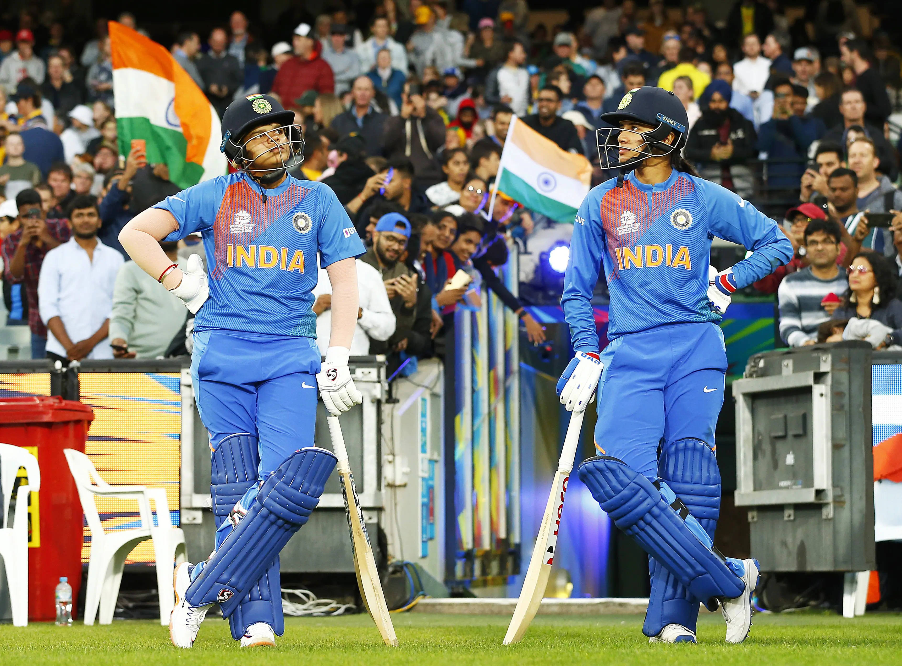 Smriti Mandhana 桌布 壁紙 — 兩名身穿藍色球衣的印度女子板球運動員自信地站在綠色球場上，手持球棒，背景中球迷揮舞著印度國旗。
