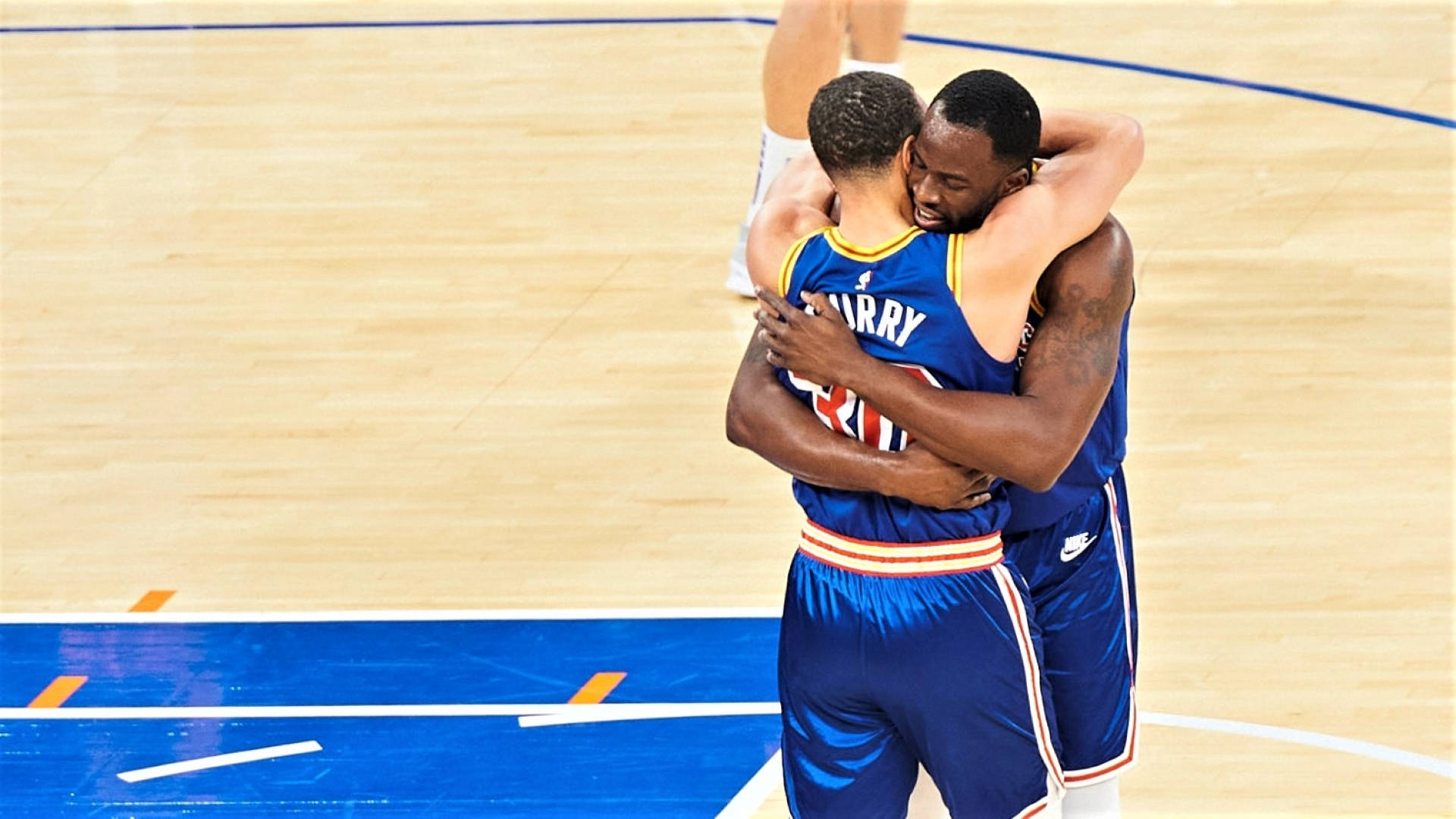Draymond Green 桌布 壁紙 — 兩名身穿藍色球衣的籃球運動員在光亮的木地板上，在明亮的場館燈光下緊緊擁抱。