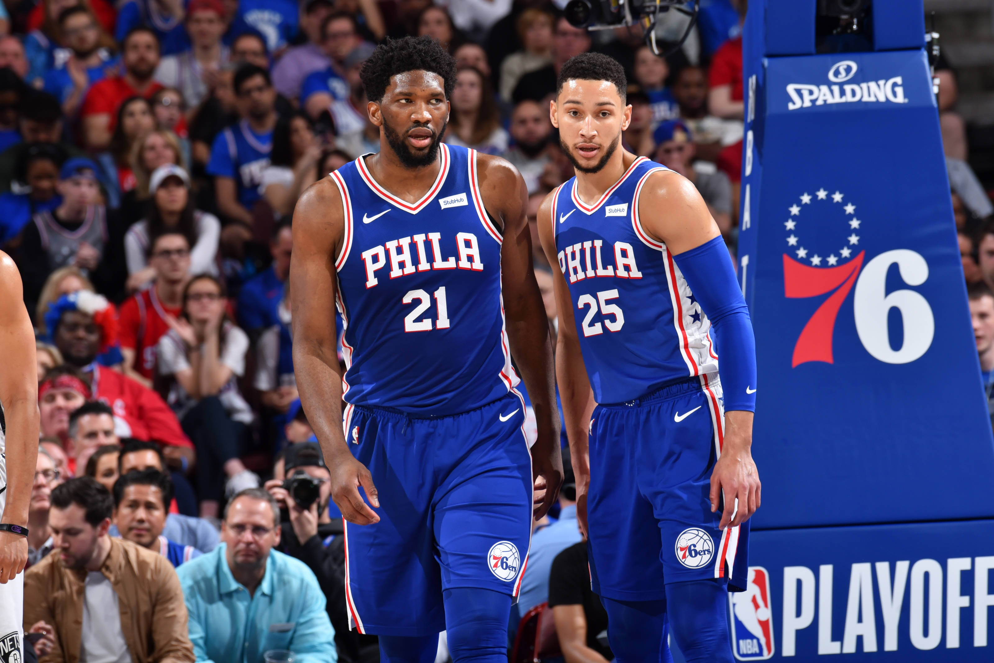 76ers 桌布 壁紙 — 兩名身穿藍色球衣的費城76人籃球運動員在季後賽比賽中站在球場上，身後有觀眾。