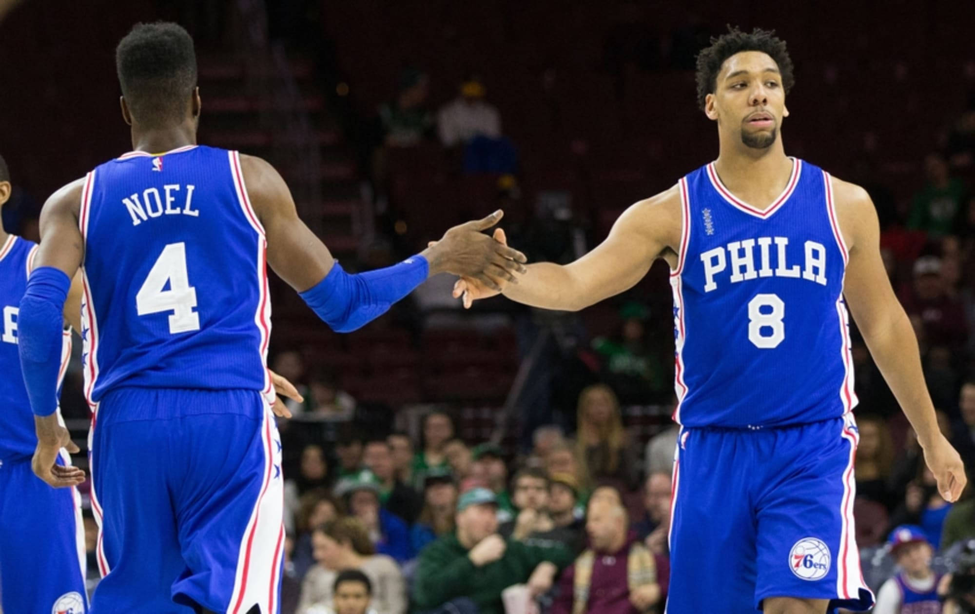 76ers 桌布 壁紙 — 兩名身穿藍色球衣的費城76人籃球運動員在燈光明亮的球場上握手，背景是模糊的觀眾。