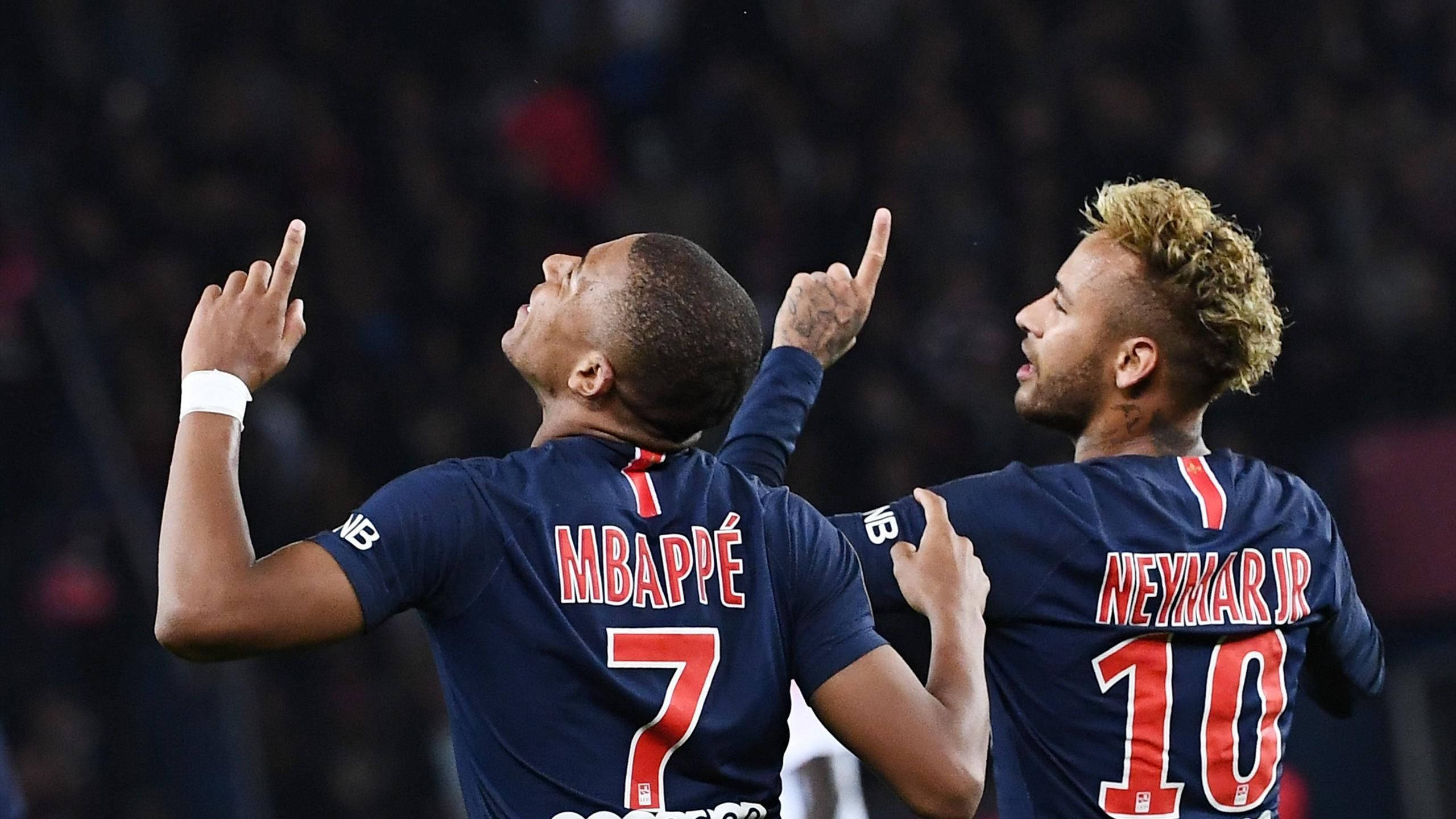 Kylian Mbappe 桌布 壁紙 — 兩名身穿藍色球衣的足球員在模糊的球場人群中舉起食指慶祝。