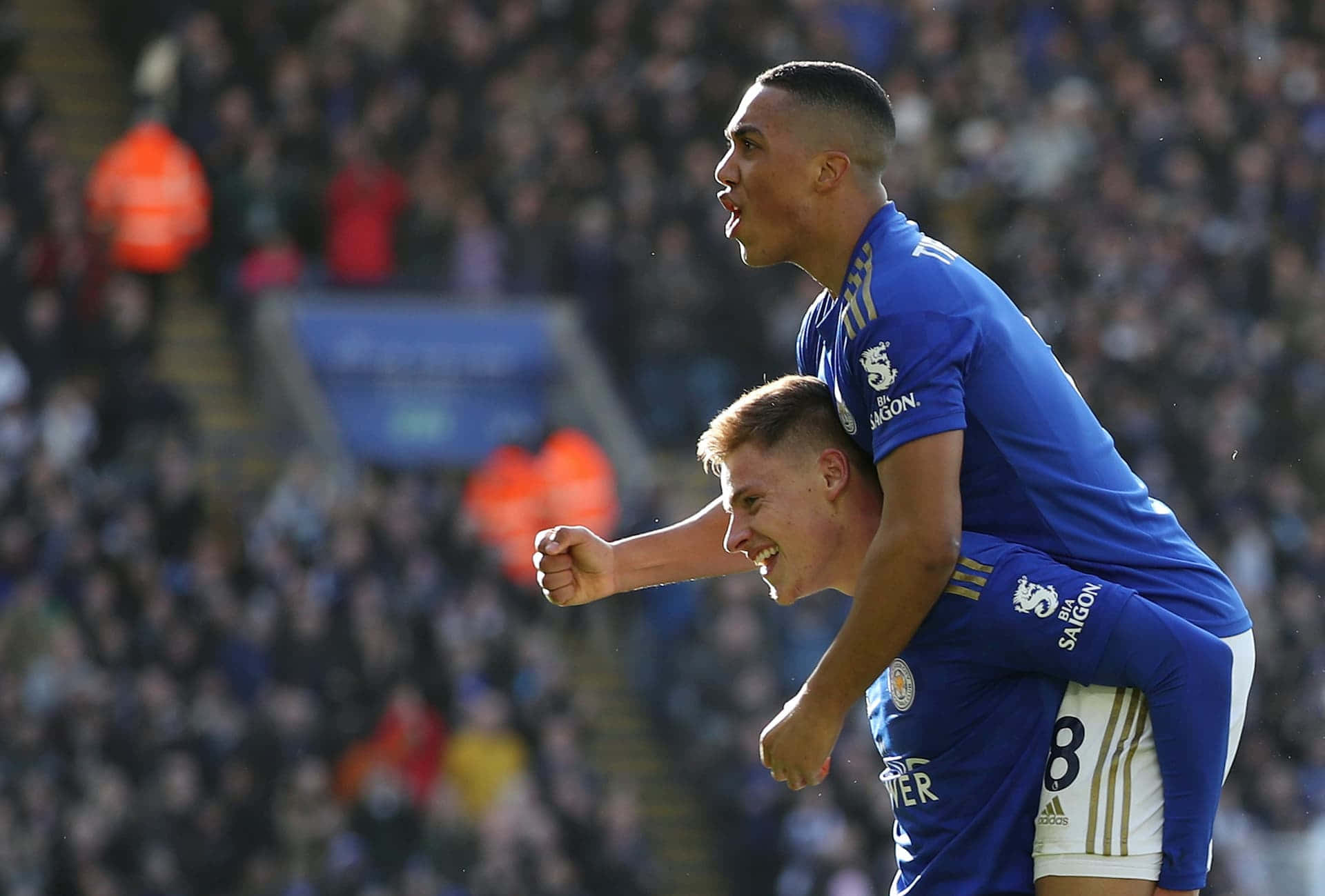 運動 桌布 壁紙 — 兩名身穿藍色球衣的足球員在陽光普照的球場人群中慶祝進球，其中一人騎在另一人背上。