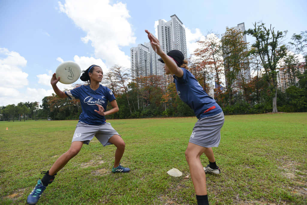 運動 桌布 壁紙 — 兩名身穿藍色球衣的運動員在草地上激烈競爭，背後樹木後方有摩天大樓在晴朗天空下聳立。