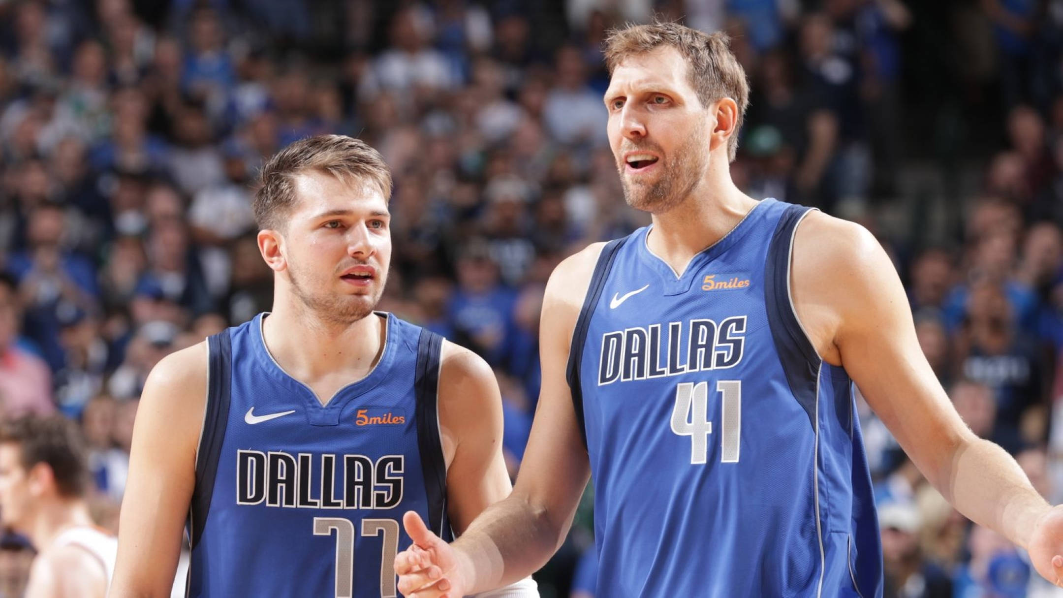 Dirk Nowitzki 桌布 壁紙 — 兩名身穿藍色球衣的達拉斯獨行俠籃球球員在比賽中互動，背後是模糊的人群。