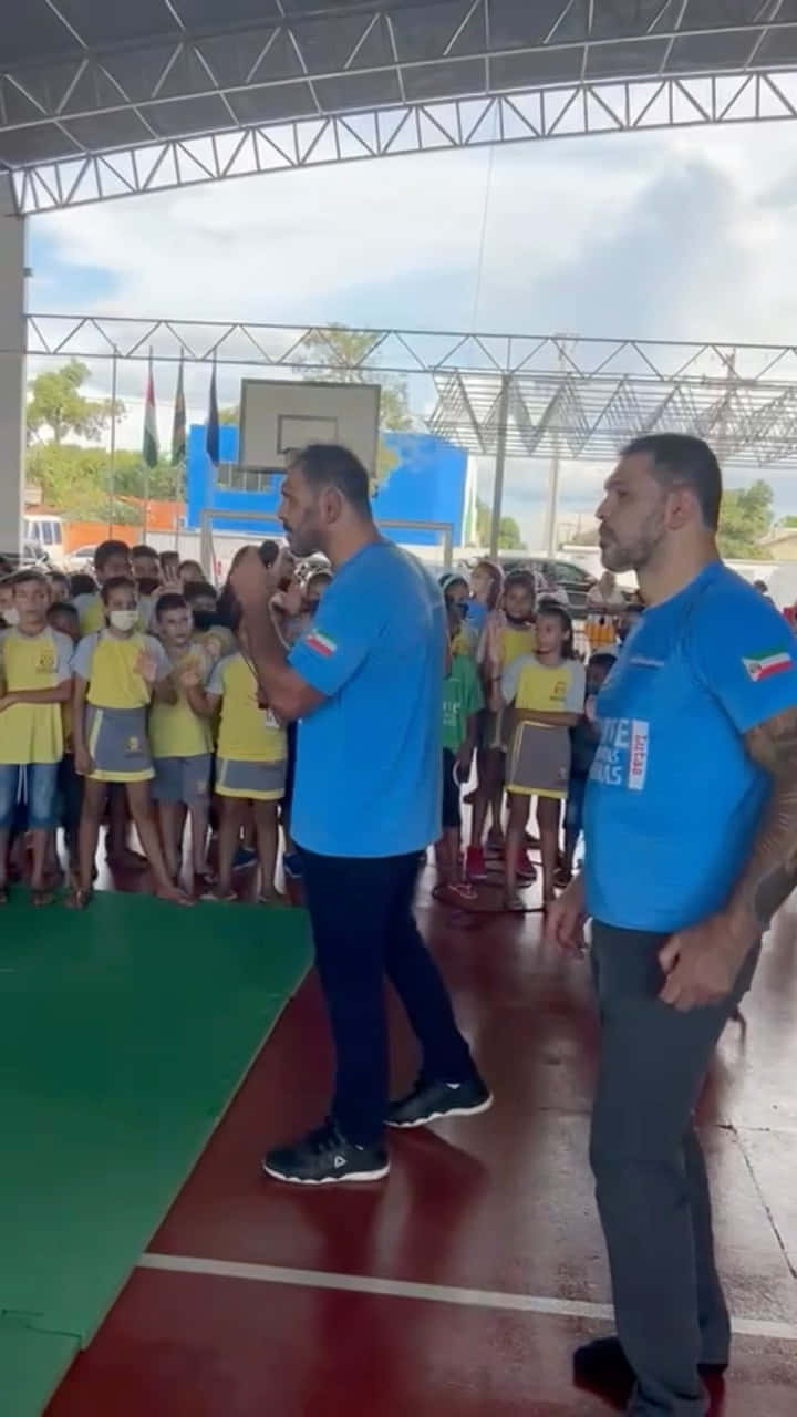 小孩動圖 桌布 壁紙 — 兩名身穿藍色襯衫的男子在一個有籃球架的遮蓋運動場下，向一群身穿黃色制服的孩子們講話。