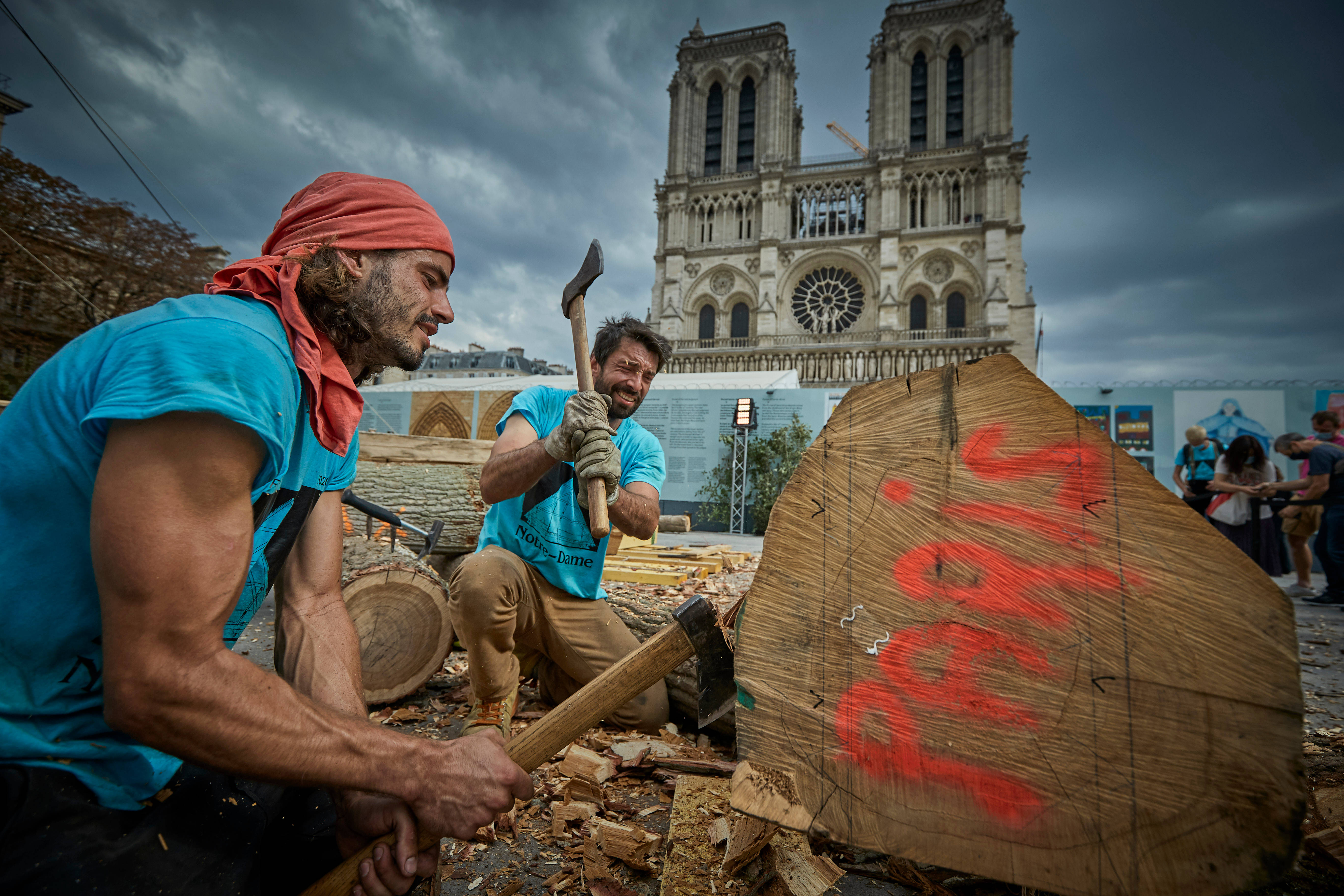 Notre Dame 桌布 壁紙 — 兩名身穿藍色襯衫的男子在暴風雨的天空下，在聖母院大教堂前用斧頭劈柴。