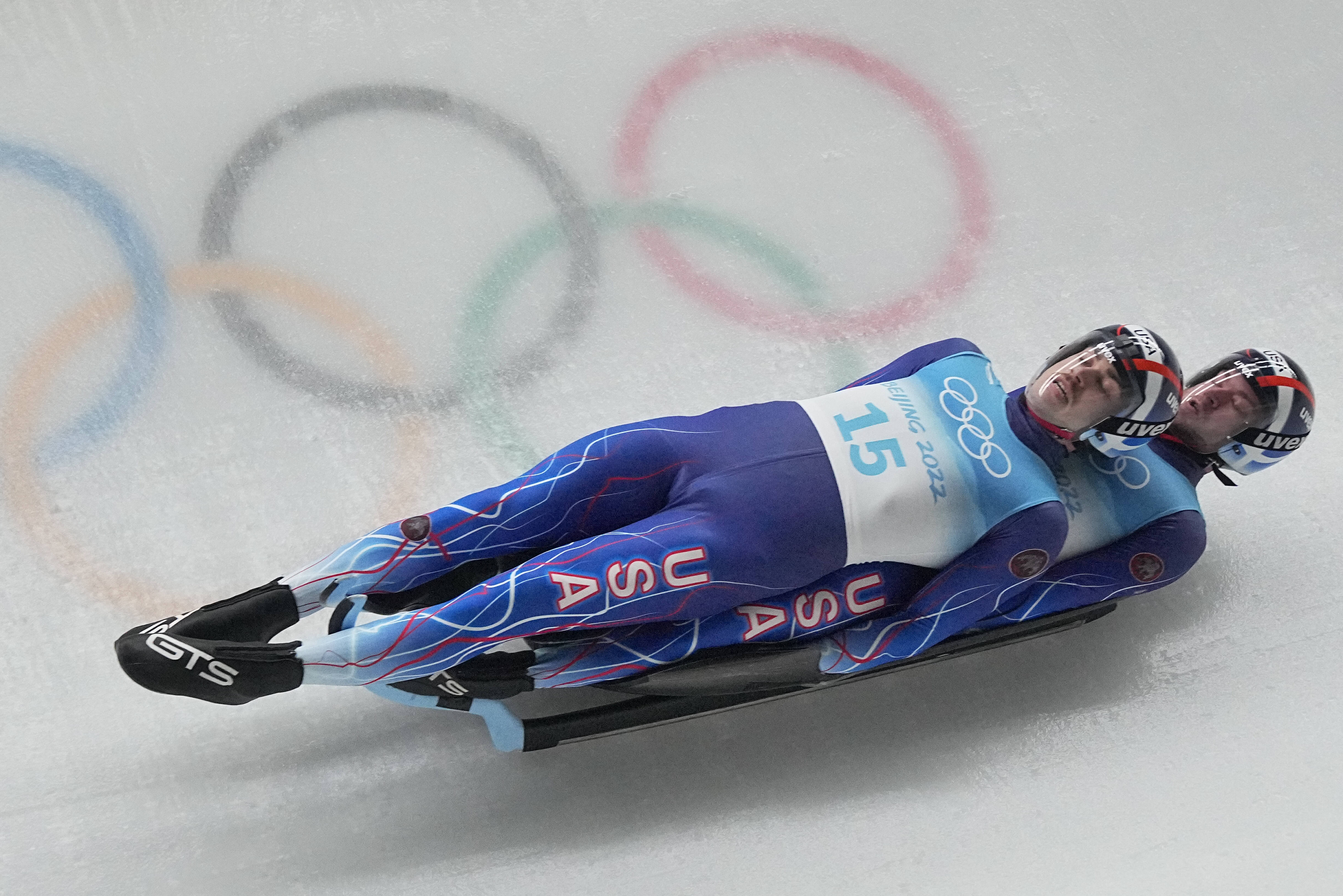 Athletic 桌布 壁紙 — 兩名身穿藍色賽車服的美國雪橇運動員在冰道上飛馳，背景中奧運五環模糊不清。
