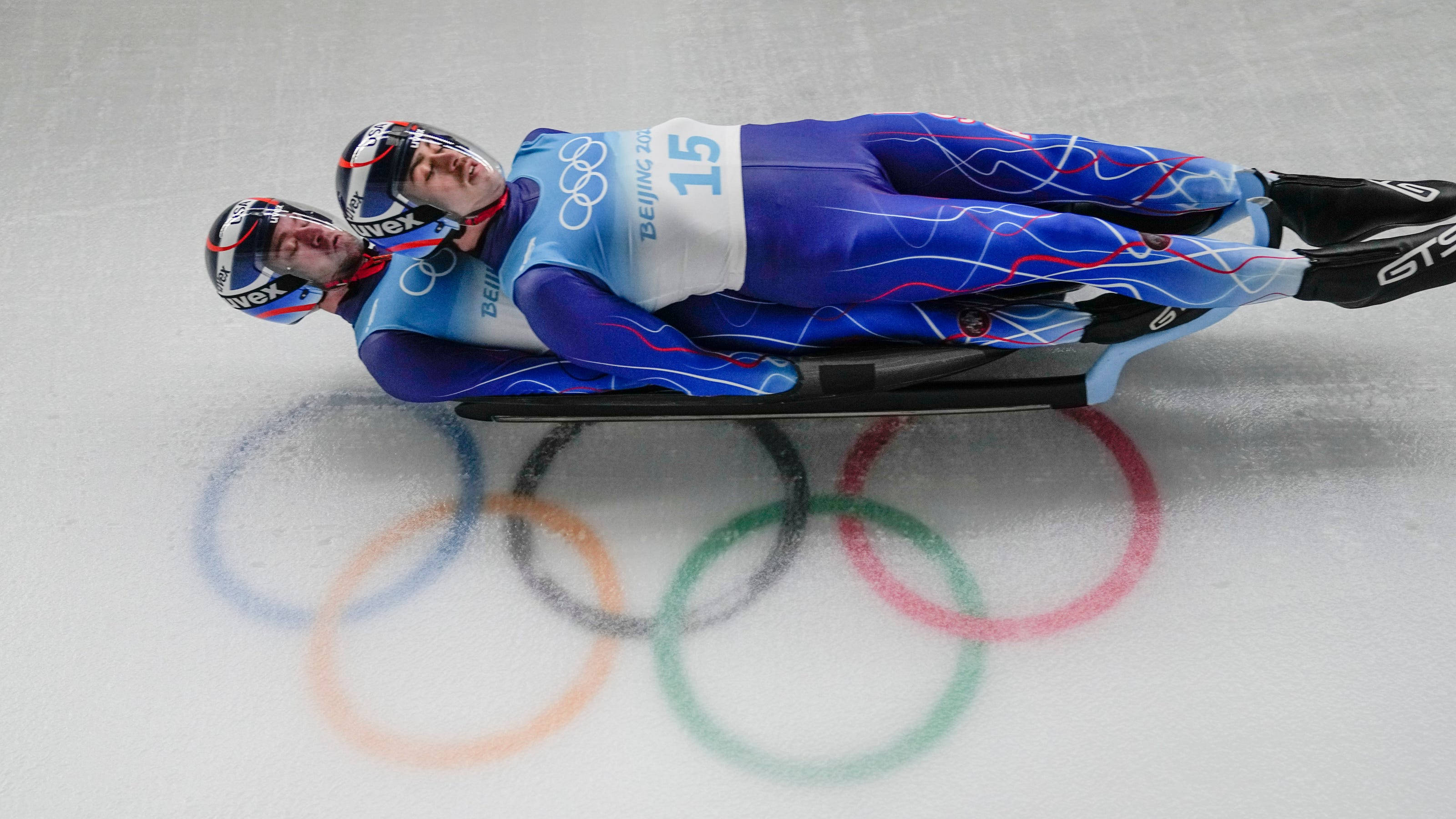 Olympics 桌布 壁紙 — 兩名身穿藍色賽車服的雪橇運動員在冰上滑行，經過彩繪的奧運五環。
