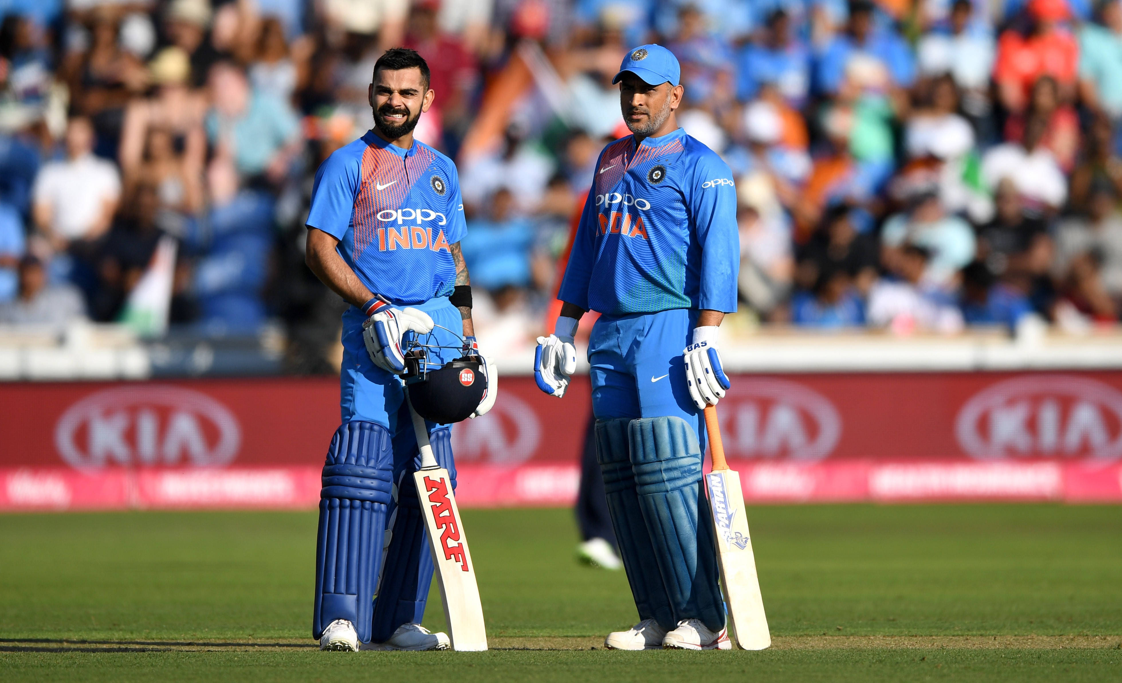 MS Dhoni 桌布 壁紙 — 兩名身穿藍色隊服的印度板球運動員站在綠色球場上，手持球棒和頭盔，身後是模糊的人群。