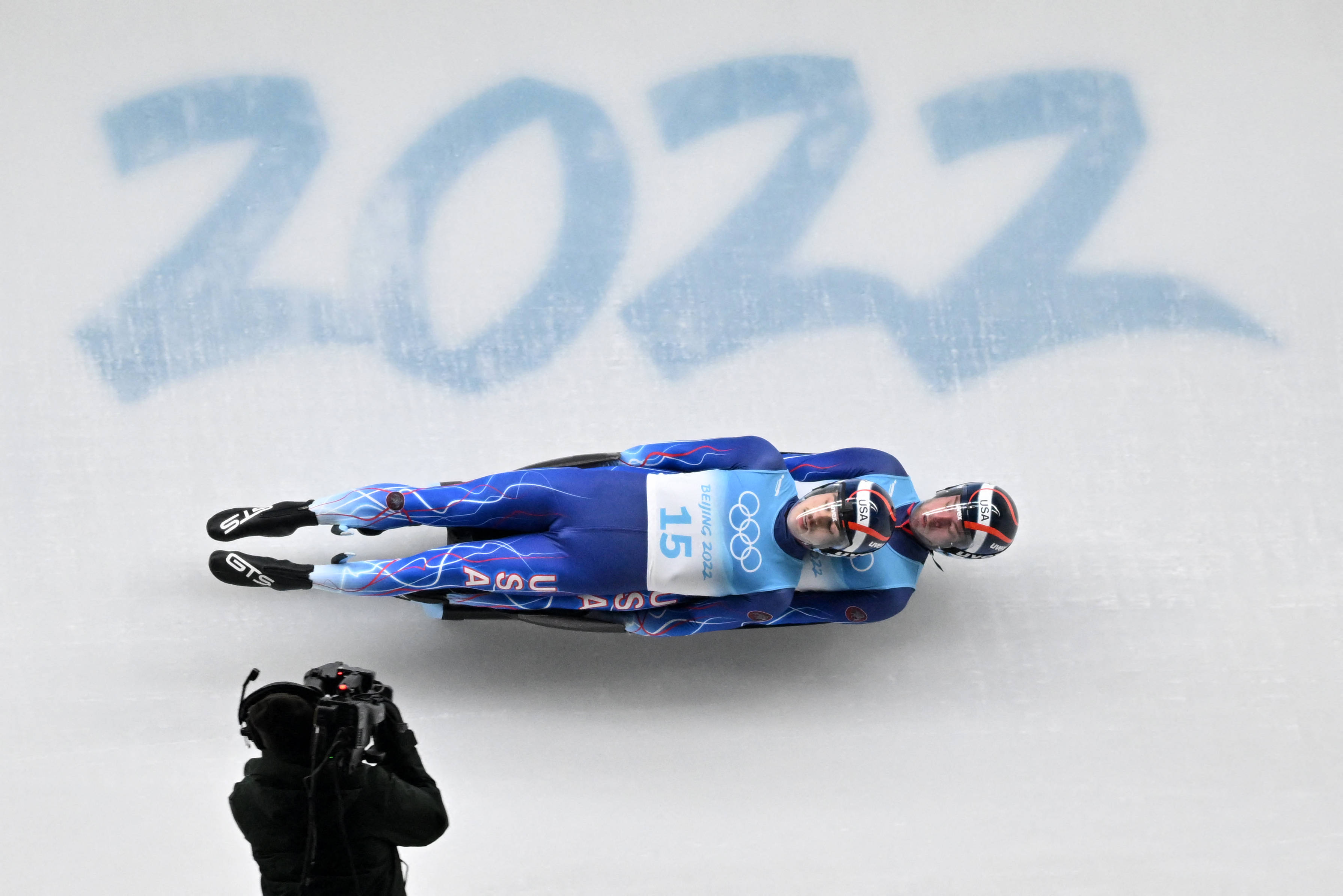 Olympics 桌布 壁紙 — 兩名身穿藍色雪橇服的運動員在冰道上競速，身後刻有「2022」，攝影師捕捉了這一刻。