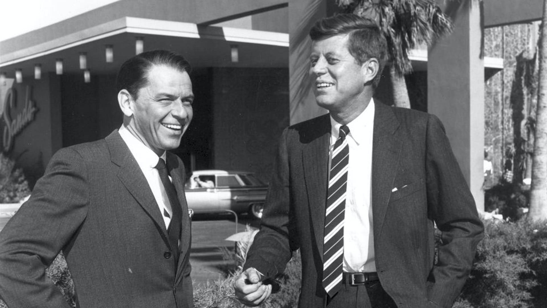 Sinatra 桌布 壁紙 — 兩名身穿西裝、面帶微笑的男子站在戶外，背景是一座中世紀現代建築，有棕櫚樹和一輛復古汽車。
