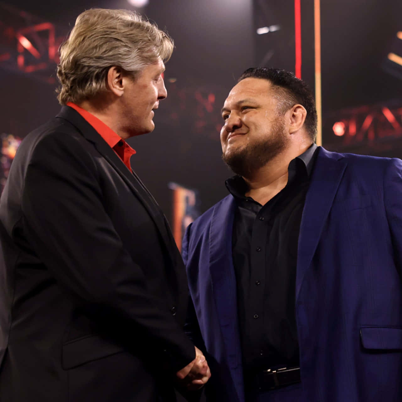 WWE 桌布 壁紙 — 兩名身穿西裝的男子在明亮的舞台燈光下握手，背景有紅色和橙色的聚光燈。