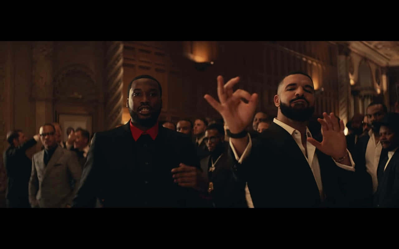 Meek 桌布 壁紙 — 兩名身穿西裝的男子在燈光昏暗、裝飾華麗的大廳中，被觀眾包圍，戲劇性地做手勢。