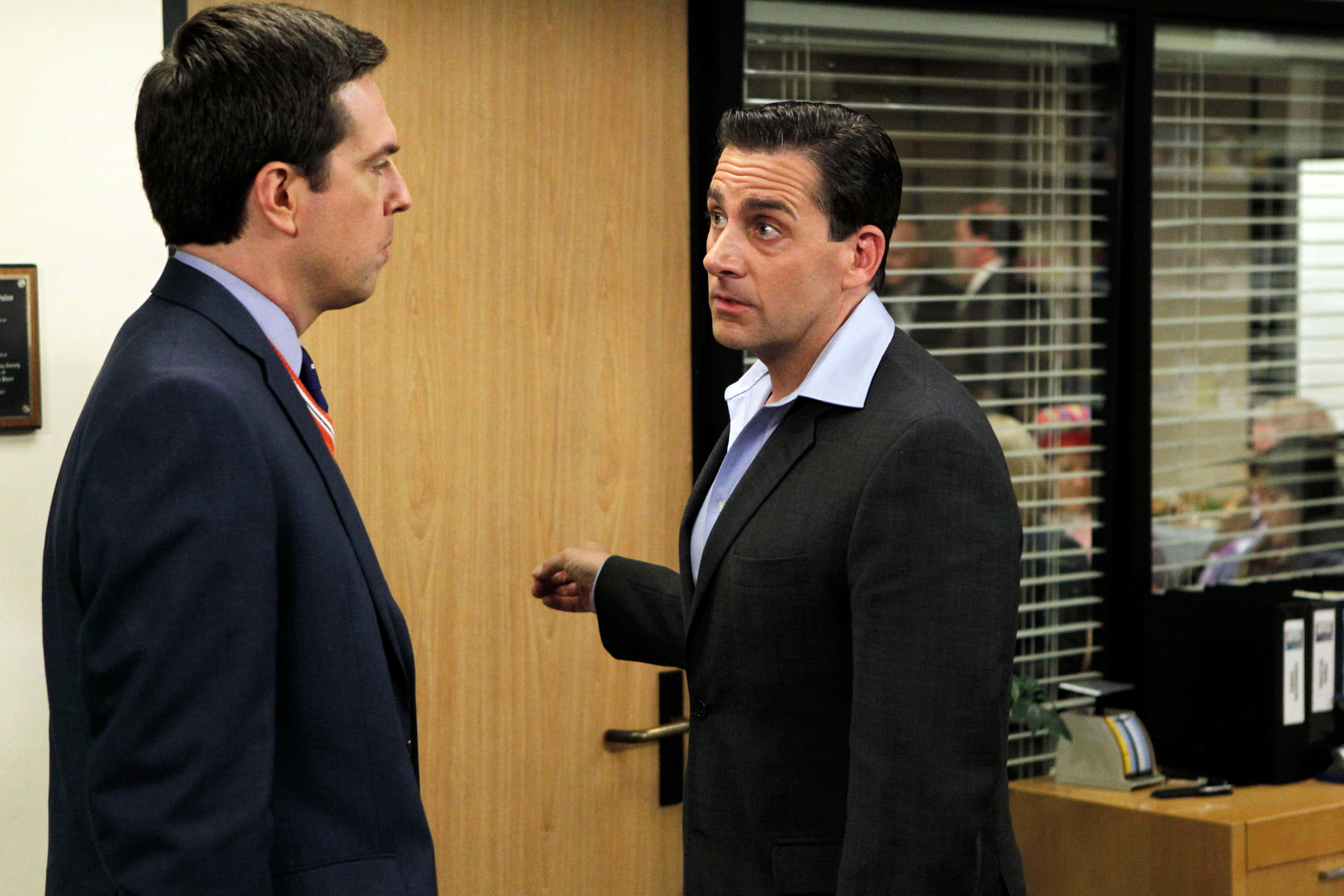 ed helms 桌布 壁紙 — 兩名身穿西裝的男子在緊張的辦公室走廊中對峙，背景有百葉窗和文件櫃。