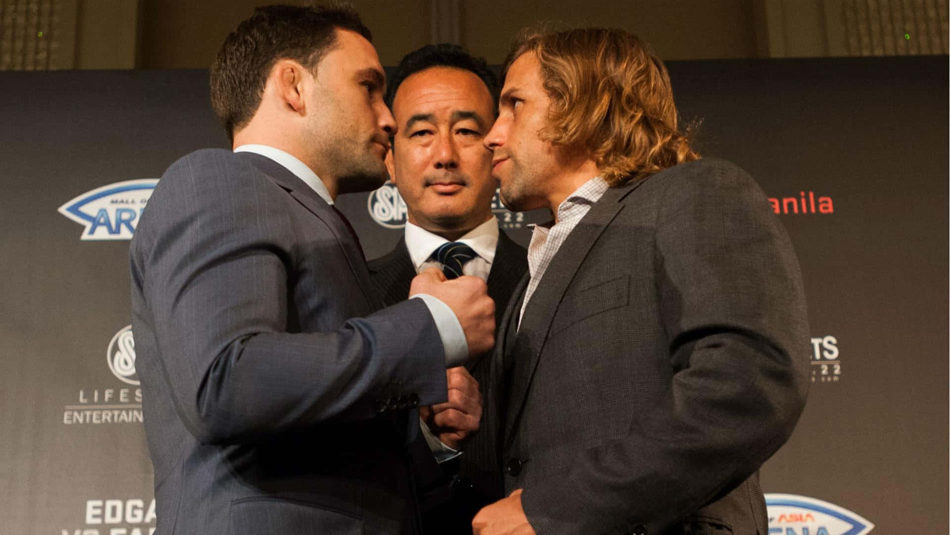 UFC 桌布 壁紙 — 兩名身穿西裝的男子在記者會上鼻對鼻對峙，第三名男子則在他們之間拿著麥克風。