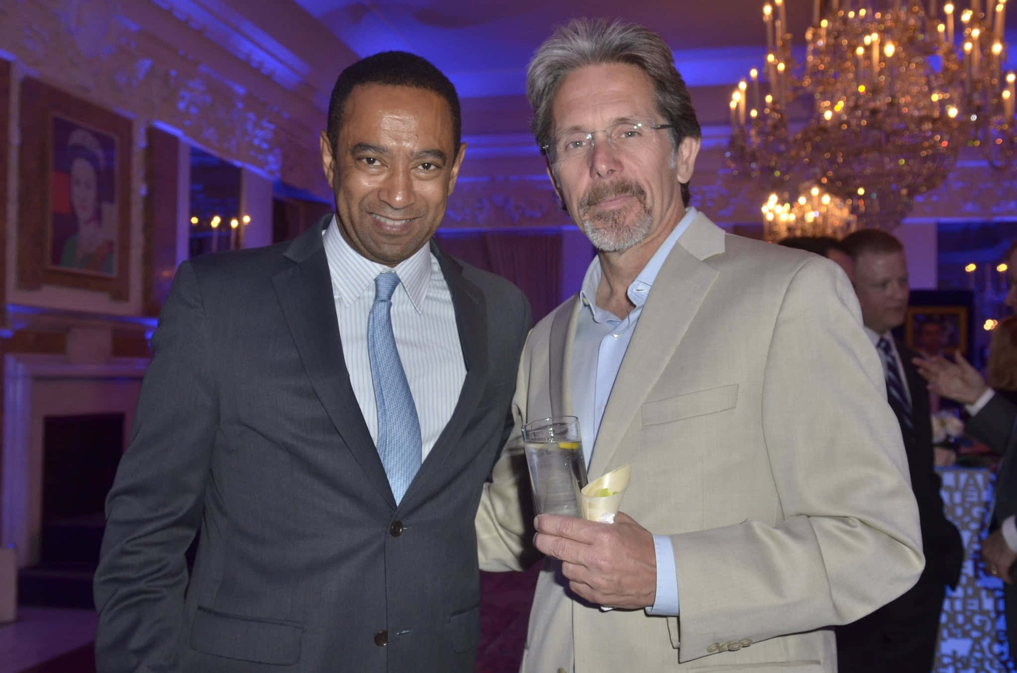 Gary Cole 桌布 壁紙 — 兩名身穿西裝的男子在閃爍的水晶吊燈下微笑拍照，背景是優雅的室內晚宴場合。