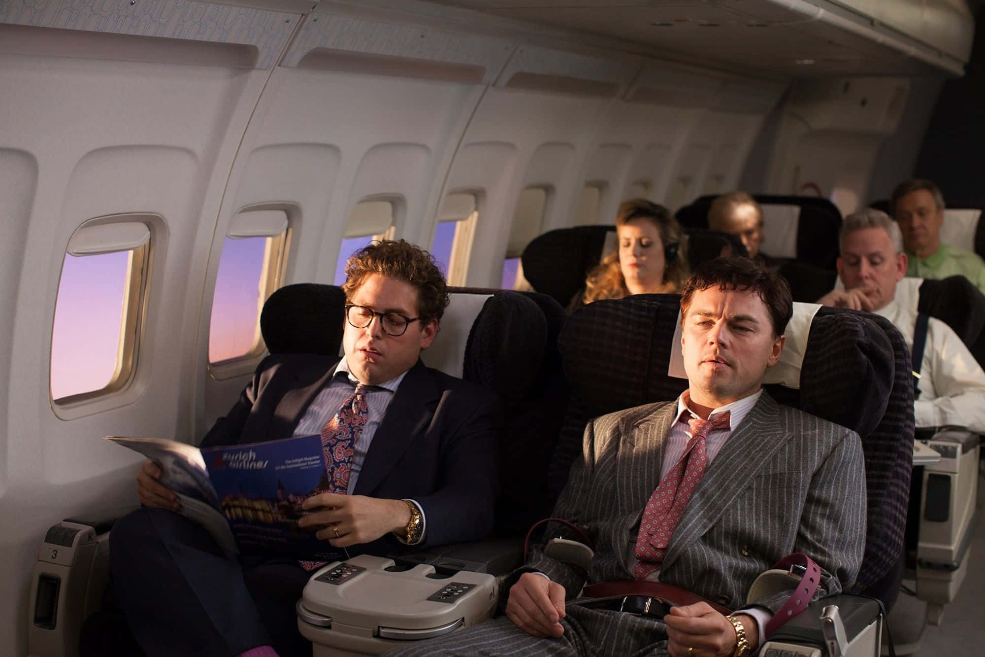 Wolf Of Wall Street 桌布 壁紙 — 兩名身穿西裝的男子坐在飛機座位上，一人正在閱讀雜誌，另一人正在睡覺，夕陽的光線從窗戶射進來。