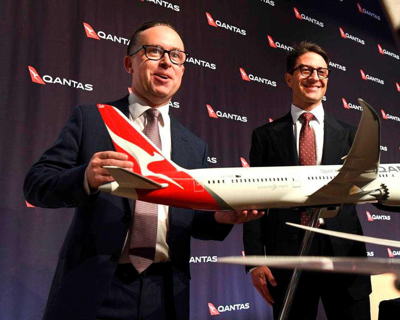  壁紙 — 兩名身穿西裝的男子微笑著，手持澳洲航空飛機模型，背景為品牌宣傳板。