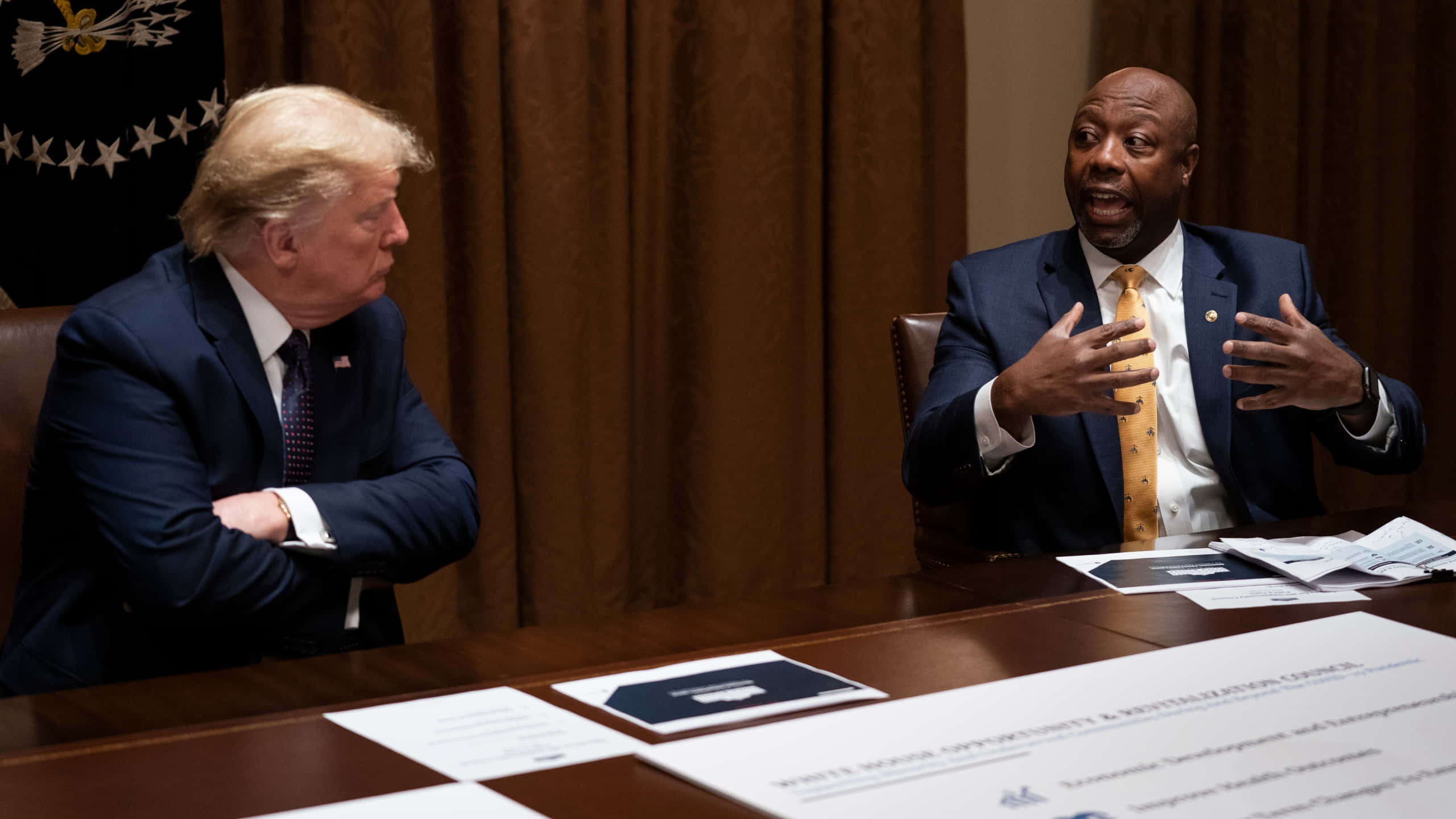Politics 桌布 壁紙 — 兩名身穿西裝的男子隔著木桌相對而坐，一人雙臂交叉聆聽，另一人則在正式會議中熱情地比劃手勢。