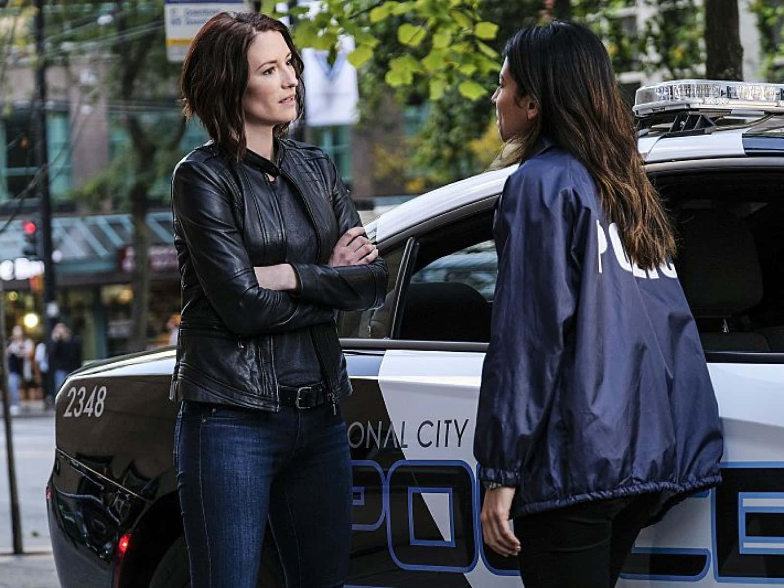 Chyler Leigh 桌布 壁紙 — 兩名身穿警察裝備的女性在城市的街道上，站在一輛有標記的巡邏車旁，面對面站立。