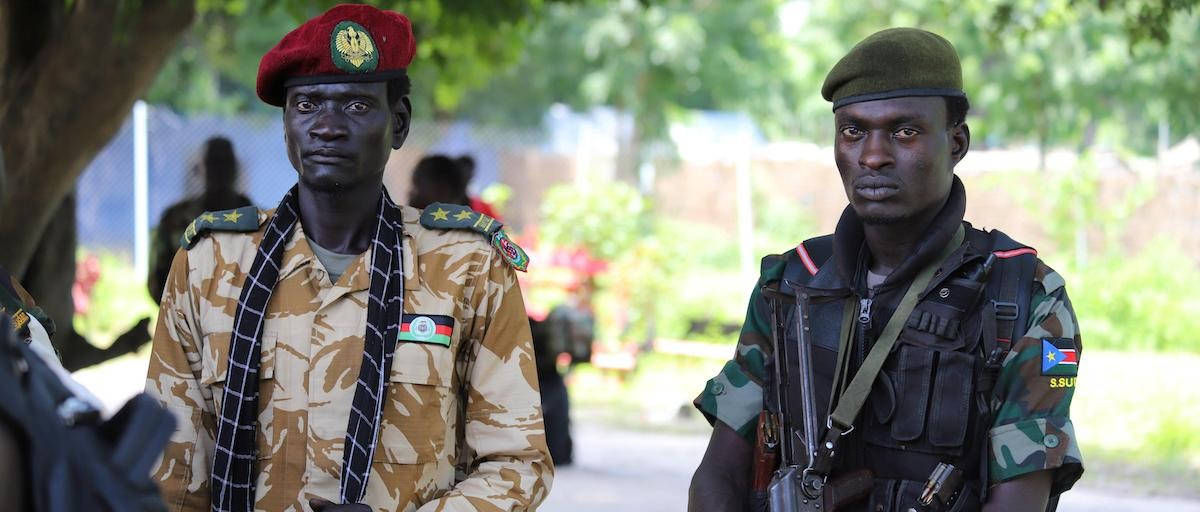 South Sudan 桌布 壁紙 — 兩名身穿迷彩服的非洲士兵在戶外警覺地站立，周圍有綠色樹葉和模糊的背景人物。