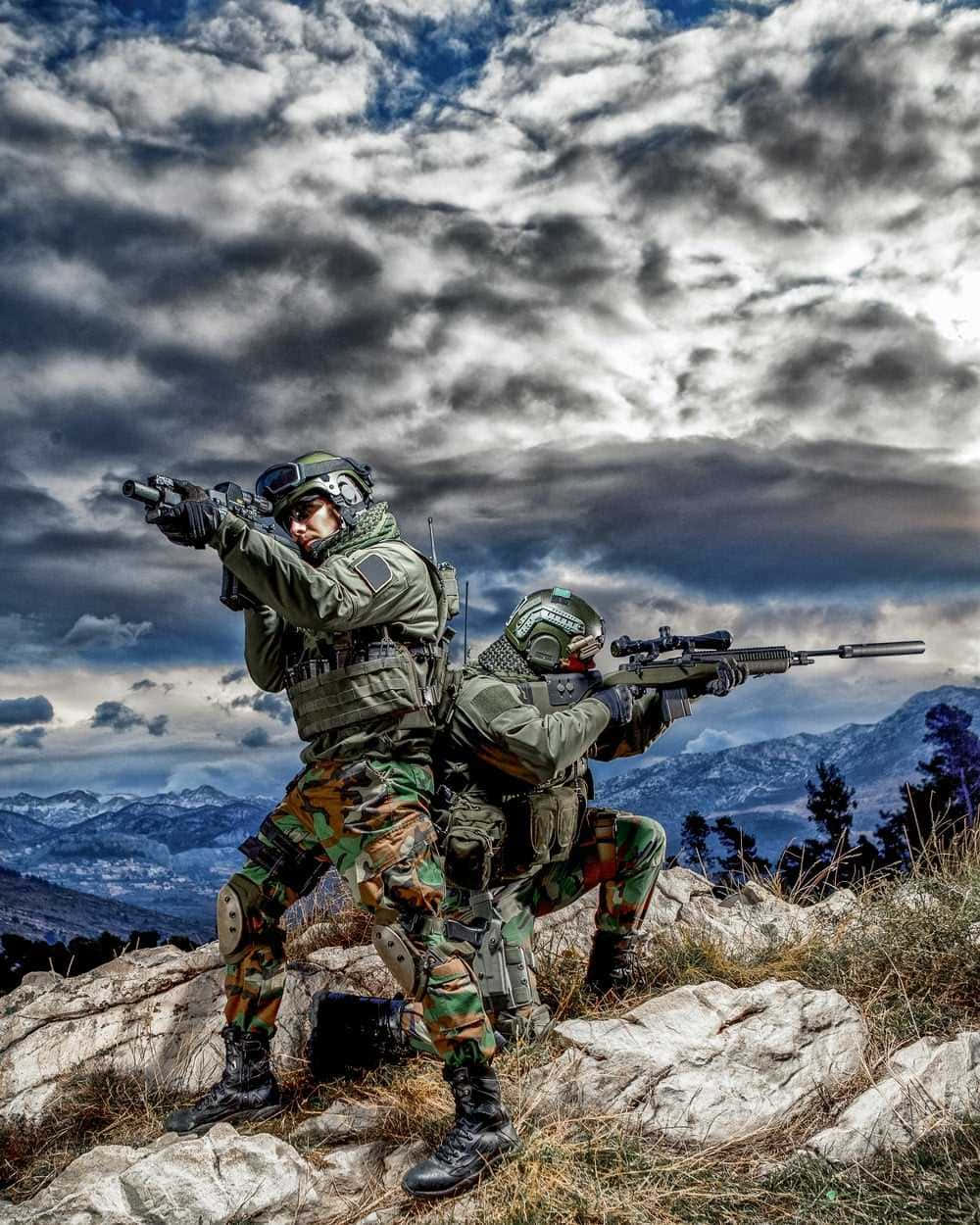 軍事動圖 桌布 壁紙 — 兩名身穿迷彩裝備的士兵在戲劇性的多雲天空下，從岩石山脊上瞄準步槍。