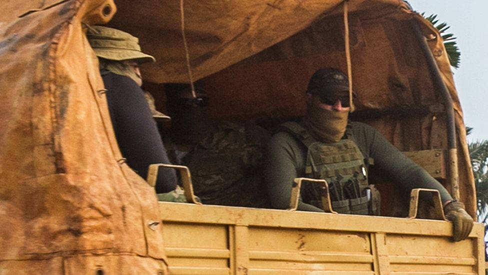 非洲 桌布 壁紙 — 兩名身穿迷彩裝備的武裝士兵在軍用卡車的開放式車斗中，在防水布下警覺地坐著。