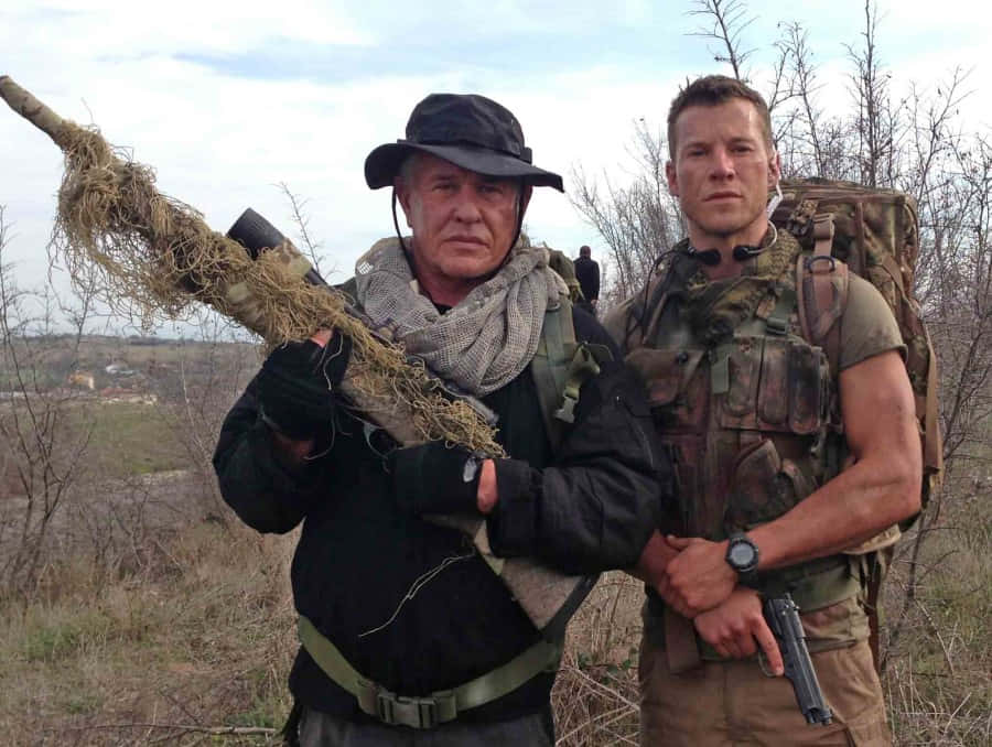 Tom Berenger 桌布 壁紙 — 兩名身穿迷彩裝備的男子站在戶外，一人手持帶有迷彩網的步槍，另一人手持手槍，兩人均神情嚴肅。