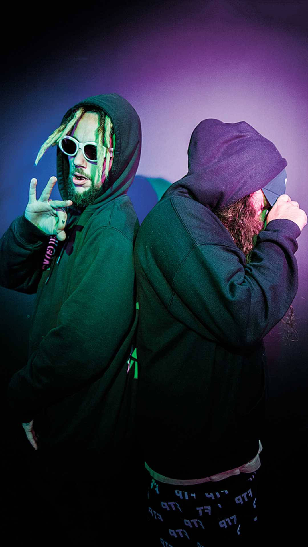 suicideboys 桌布 壁紙 — 兩名身穿連帽上衣的男子在紫色和藍色舞台燈光下背對背擺姿勢，一人做手勢，另一人調整眼鏡。