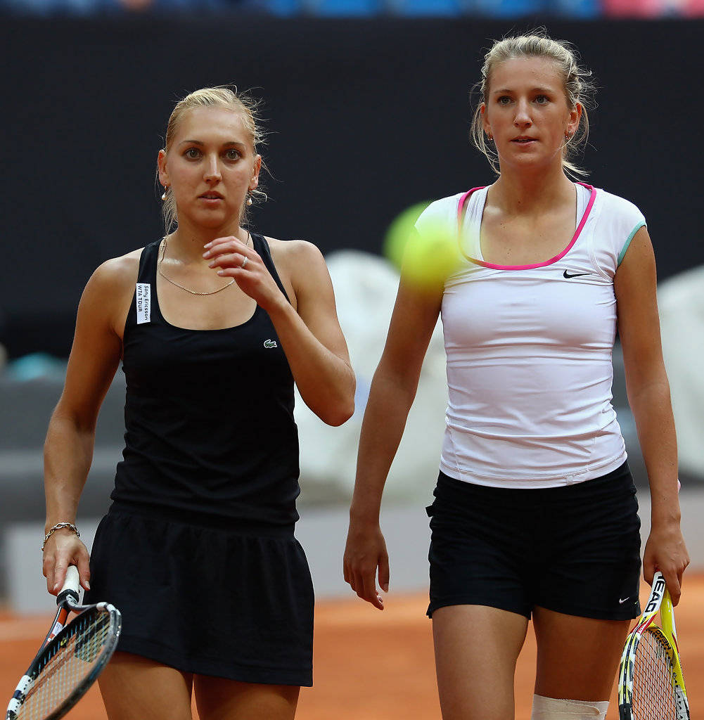 Tennis 桌布 壁紙 — 兩名身穿運動裝備的女網球選手站在紅土球場上，一個模糊的網球在她們之間移動。
