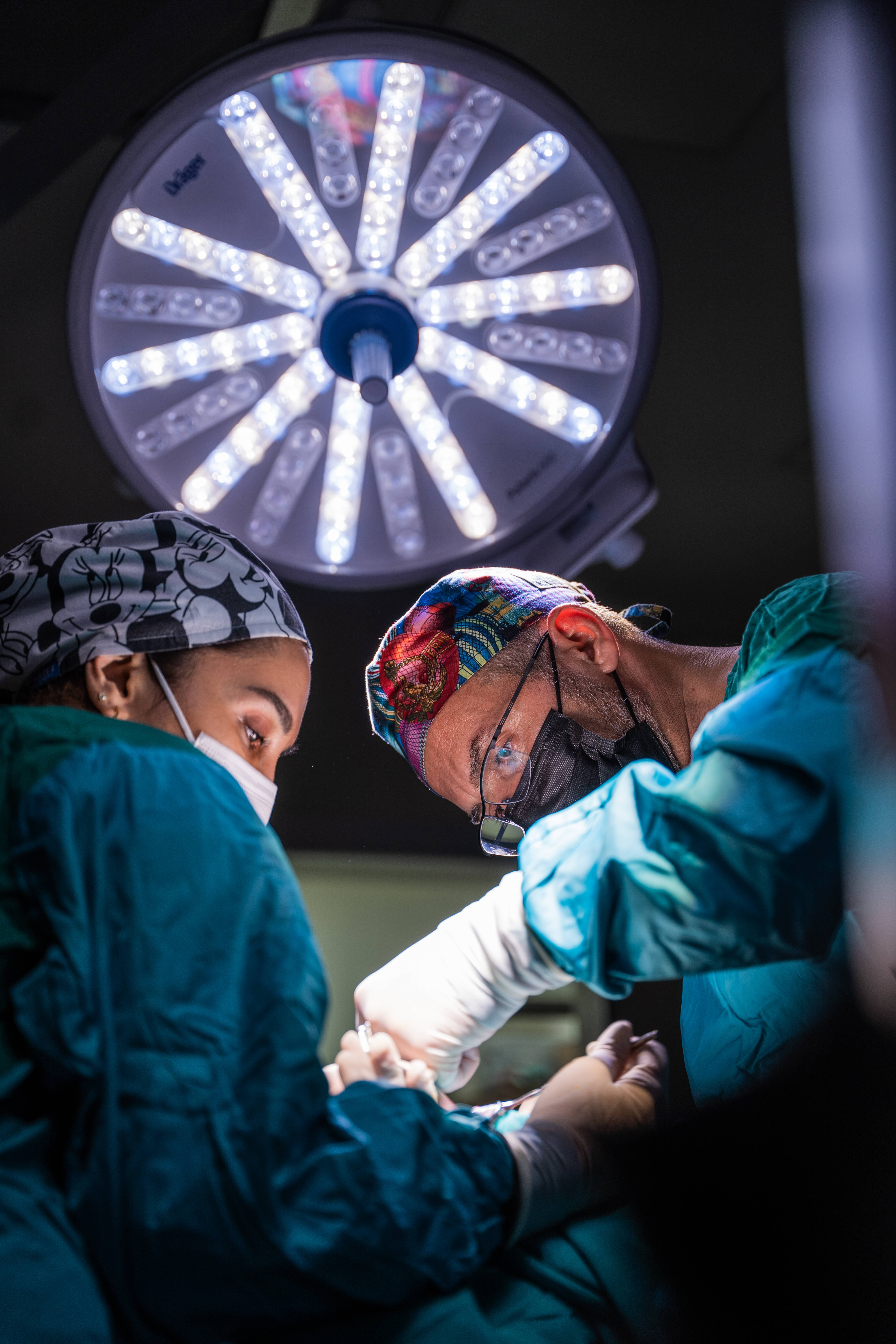 Doctors 桌布 壁紙 — 兩名身穿青綠色手術服和圖案帽子的外科醫生在昏暗的手術室中，在明亮的手術燈下專注地進行手術。