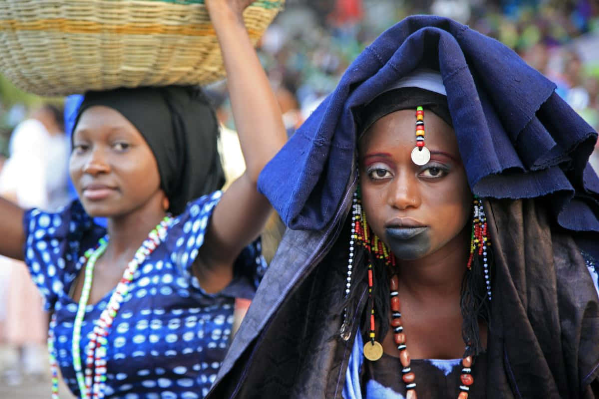 African Culture 桌布 壁紙 — 兩名身穿鮮豔傳統服飾的女性在熱鬧的文化聚會中頭頂編織籃子。