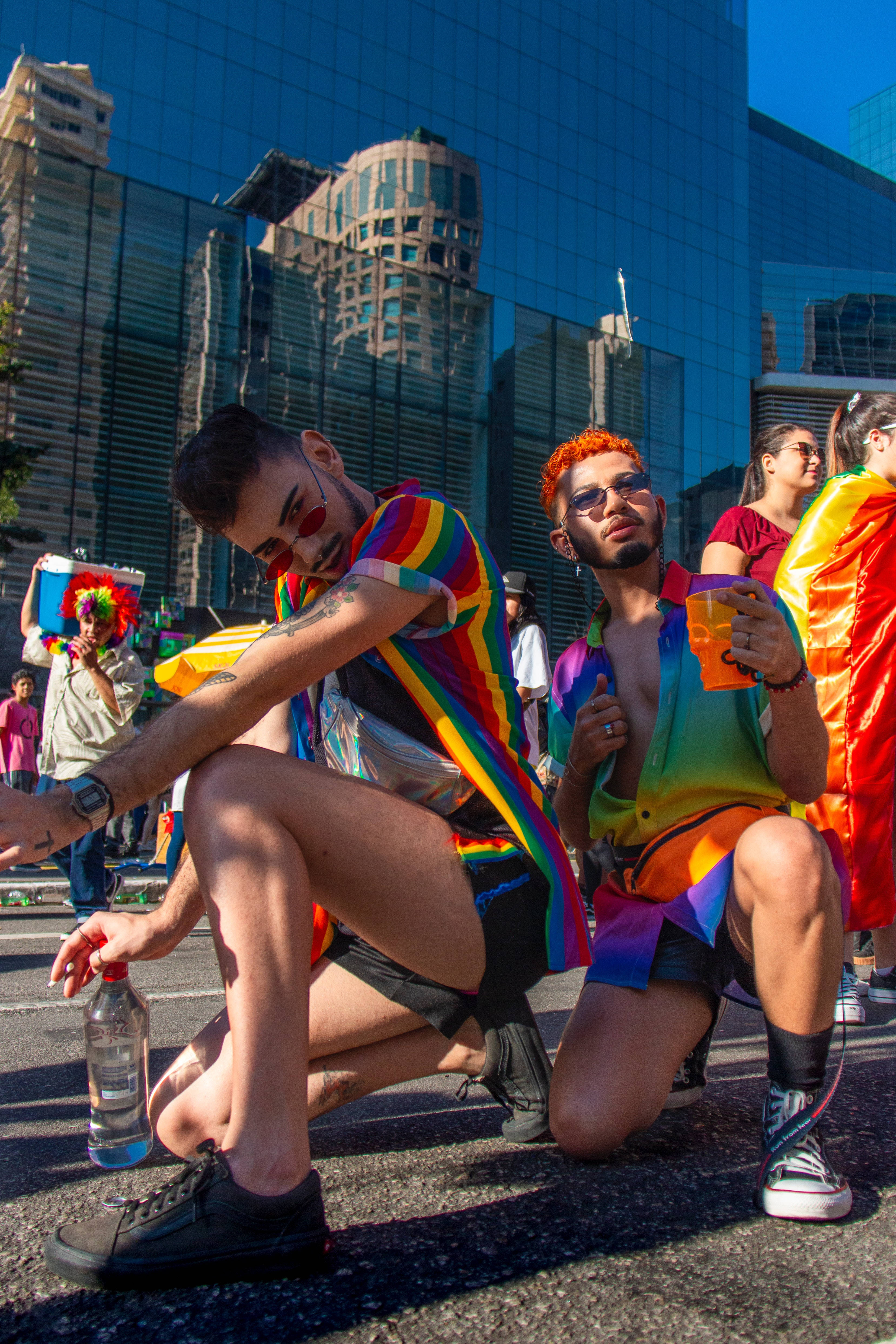 twink 桌布 壁紙 — 兩名身穿鮮豔彩虹服裝的男子在陽光明媚的遊行中蹲在城市的街道上，背景中摩天大樓倒映著。