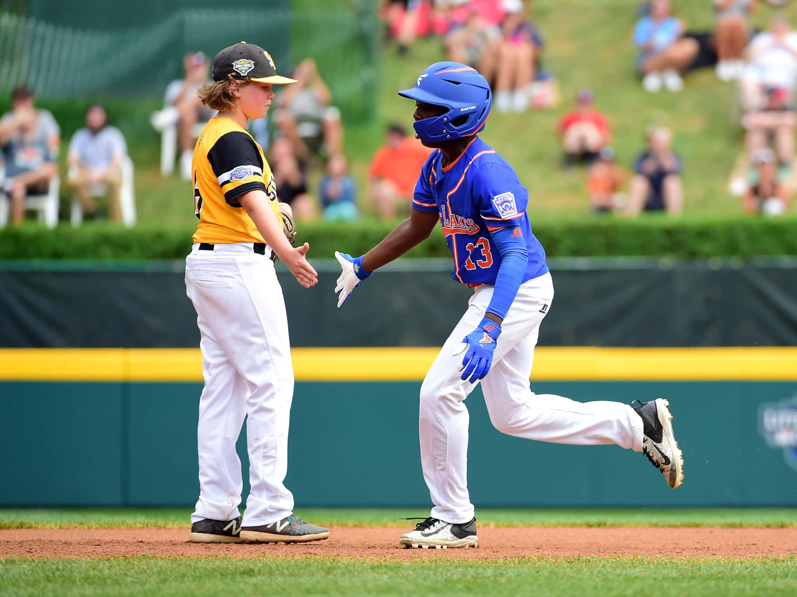 Little League Baseball 桌布 壁紙 — 兩名身穿鮮豔球衣的年輕棒球運動員在陽光普照的比賽中擊掌，背景有觀眾。