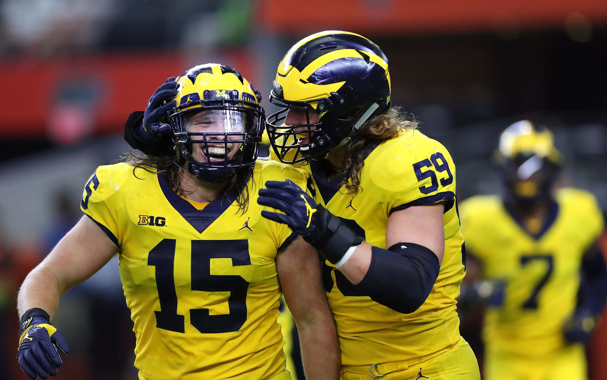 Football Players 桌布 壁紙 — 兩名身穿黃色球衣的密西根大學美式足球員在夜間比賽中於球場上激烈慶祝。