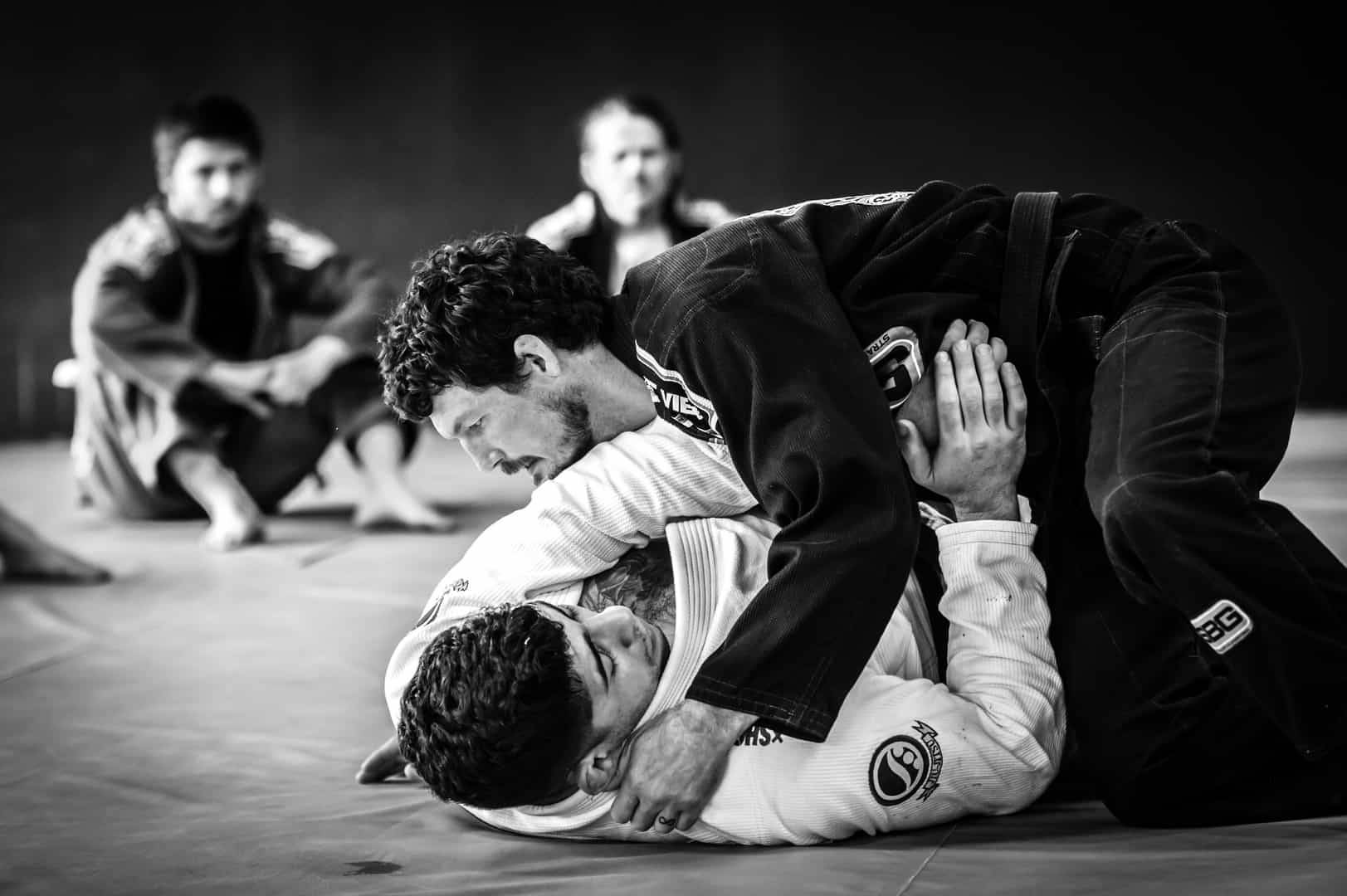 martial arts 桌布 壁紙 — 兩名身穿黑白道服的男子在軟墊上激烈纏鬥，背景有兩個模糊的觀察者。