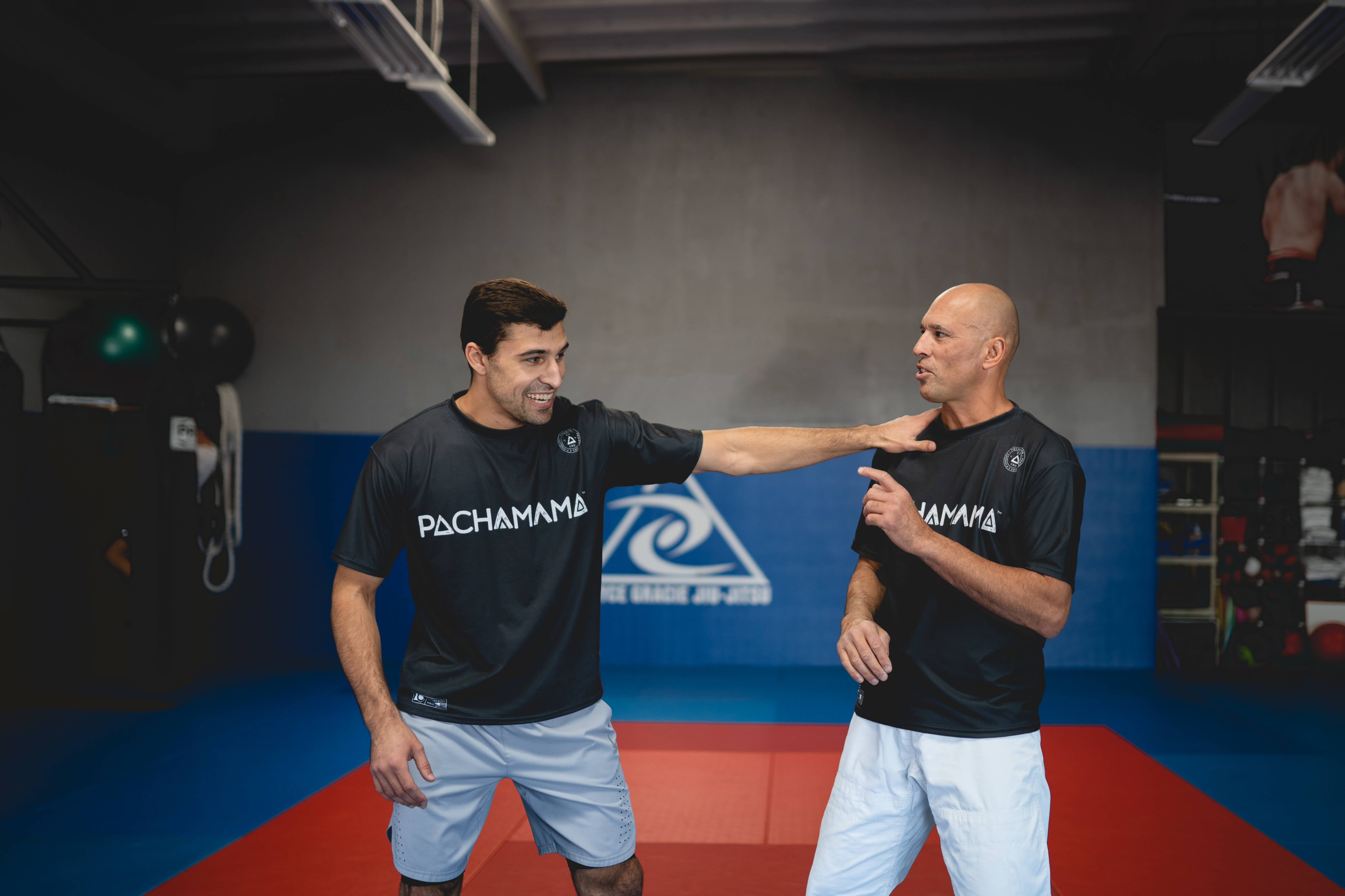 MMA 桌布 壁紙 — 兩名身穿黑色「PACHAMAMA」襯衫的男子在光線昏暗的健身房的紅藍色墊子上進行專注的武術訓練。